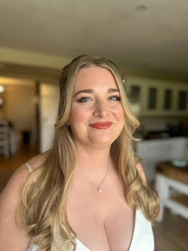 A young woman with long blonde hair and blue eyes smiling indoors, wearing a white top and a delicate necklace.