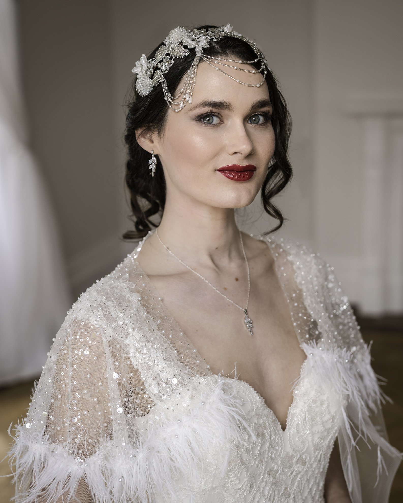 Claire Field Bridal Hair and Make up. Close-up of a woman in a wedding dress, wearing a pearl and crystal headpiece and earrings, with makeup including red lipstick, and a necklace with a large pendant.