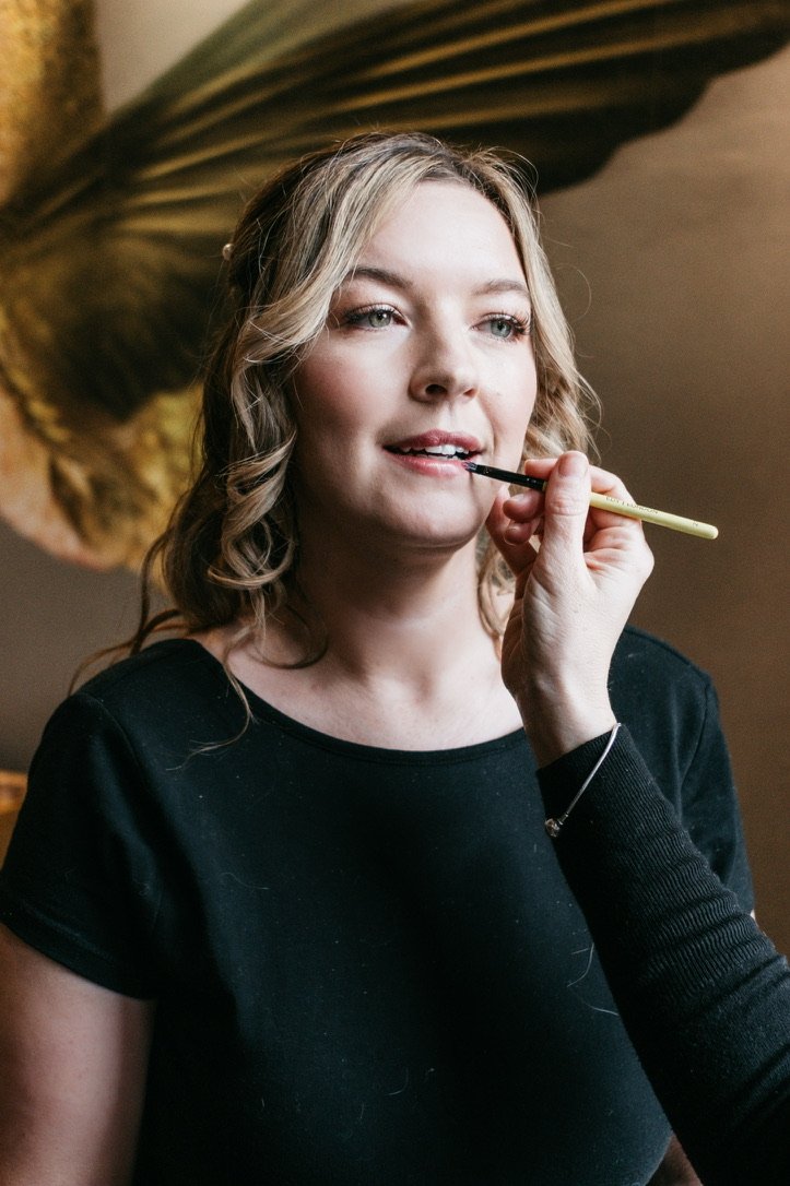 Claire Field Bridal Hair and Make up. A woman with blonde hair in loose curls is getting her makeup done, with a makeup brush near her lips. She is wearing a black top and has a large green plant in the background.