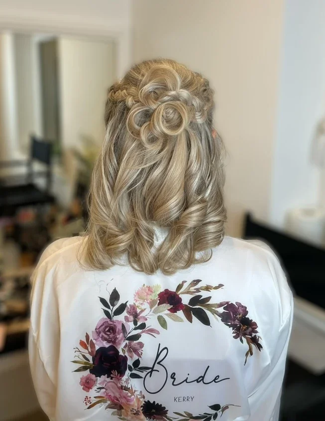 Claire Field Bridal Hair and Make up. Back view of a woman with styled blonde hair with loose curls and decorative curls on top, wearing a white robe with a floral wreath and the word 'Bride' written on it.