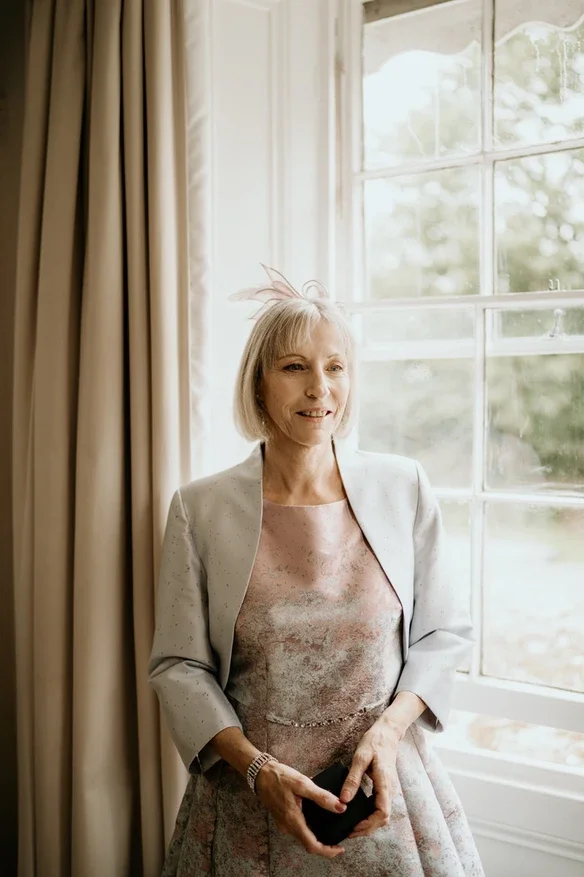 Claire Field Bridal Hair and Make up. A woman standing near a large window with curtains, holding a smartphone, smiling, wearing a light-colored suit jacket over a pastel dress with a fascinator in her hair.