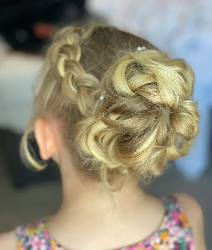Claire Field Bridal Hair and Make up. Back view of a woman with blonde hair styled in an intricate updo with curls and braids, adorned with small hair accessories, wearing a colorful floral dress.