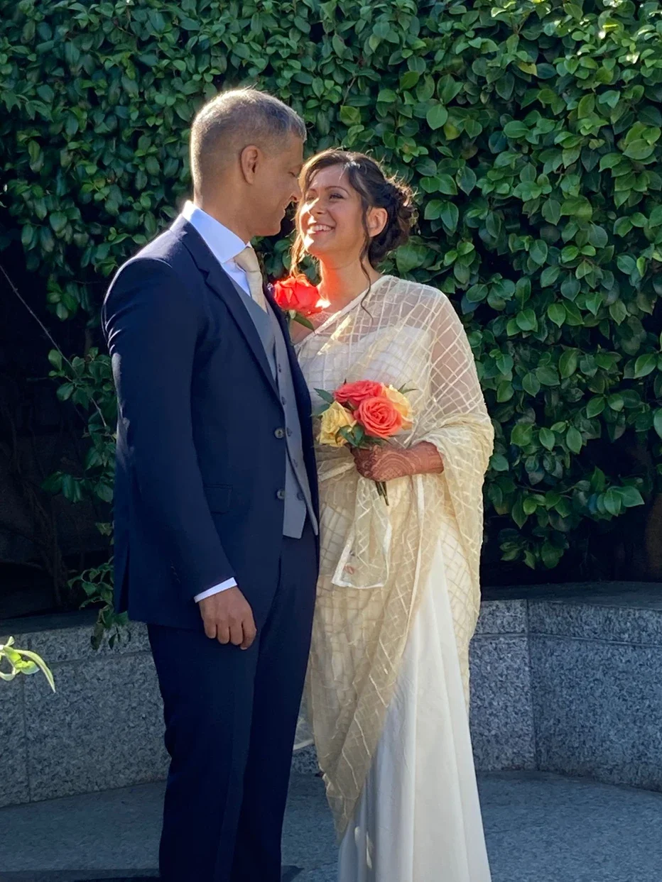 Claire Field Bridal Hair and Make up. A bride and groom standing close together outdoors on their wedding day, smiling at each other. The bride is holding a bouquet of pink and yellow roses, wearing a cream saree with lace detailing.