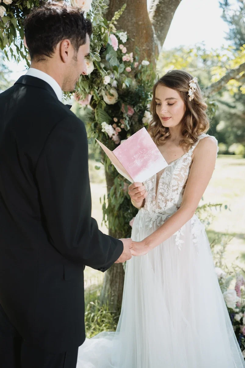 Claire Field Bridal Hair and Make up. A bride and groom hold hands during their outdoor wedding ceremony, with the bride reading vows from a pink booklet under a floral arch.