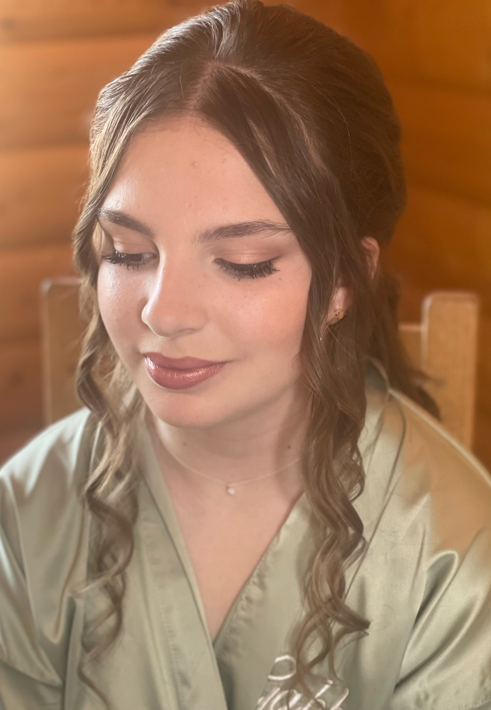 Claire Field Bridal Hair and Make up. A woman with loose curls and subtle makeup, smiling softly with her eyes closed, sitting indoors with a wooden wall in the background.