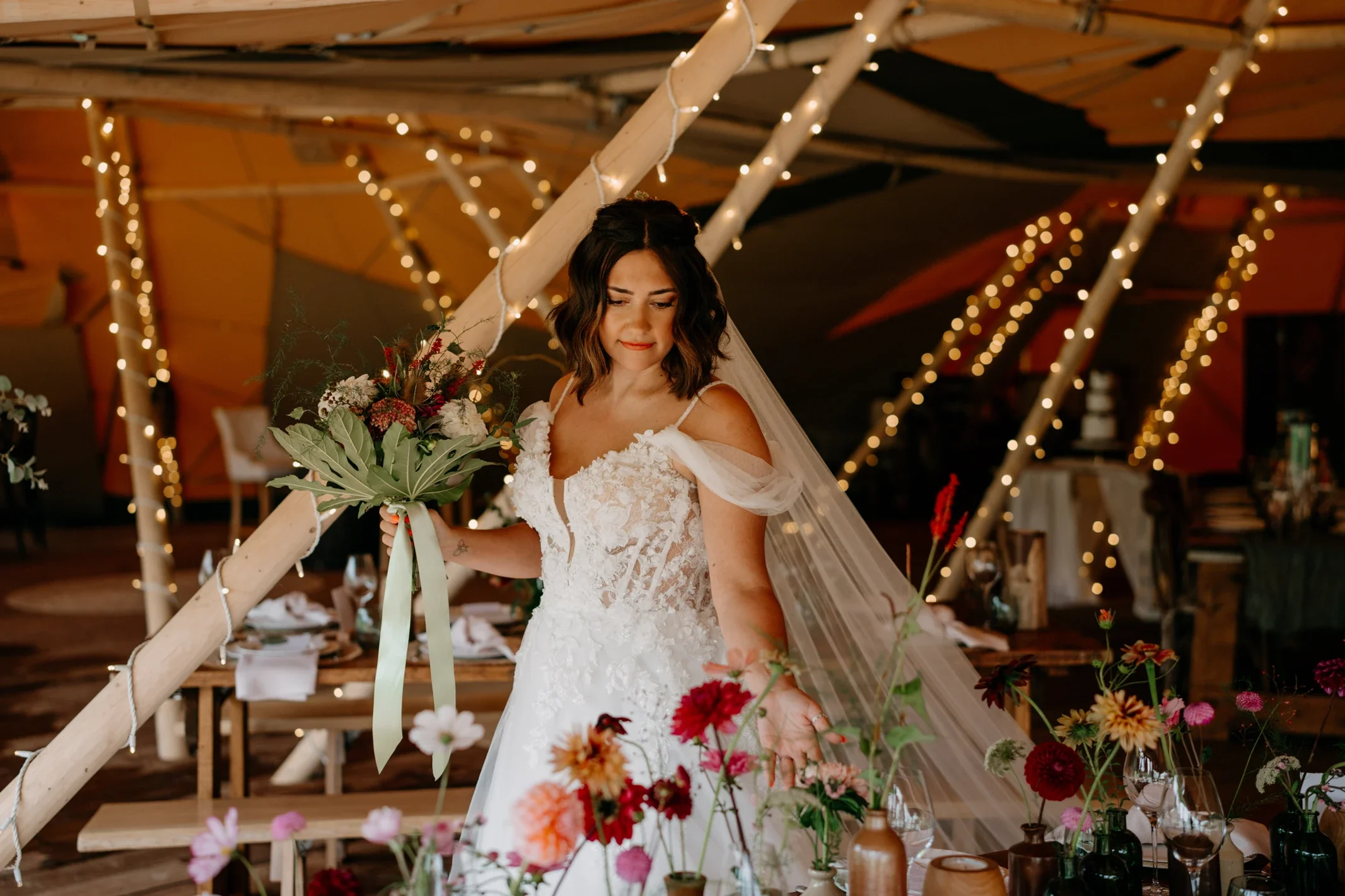 Claire Field Bridal Hair and Make up. A bride in a lace wedding dress holds a bouquet of flowers at her reception decorated with string lights and floral arrangements.