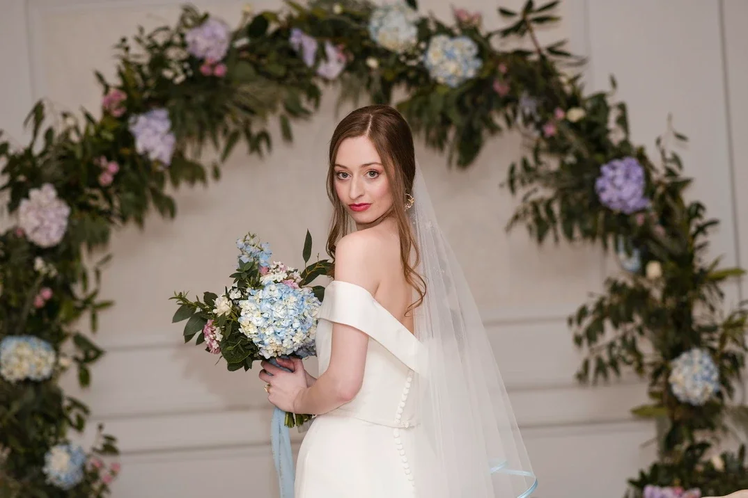 Claire Field Bridal Hair and Make up. A bride in an off-shoulder wedding dress holding a bouquet of flowers, standing in front of a floral arch with pink, purple, and white flowers and green leaves.