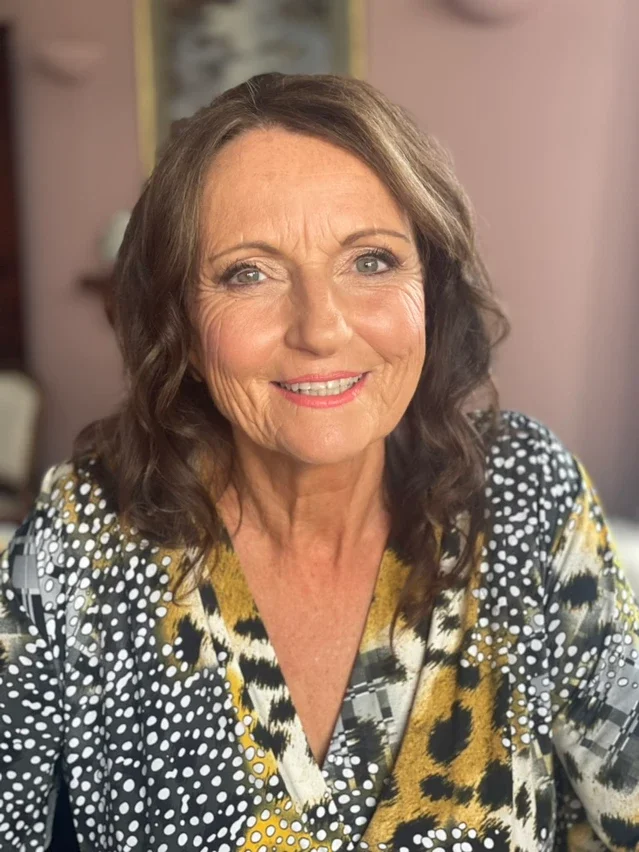 Claire Field Bridal Hair and Make up. A smiling middle-aged woman with shoulder-length brown wavy hair, wearing a patterned top with black, white, yellow, and gold colors, in a room with soft pink walls and some framed pictures in the background.
