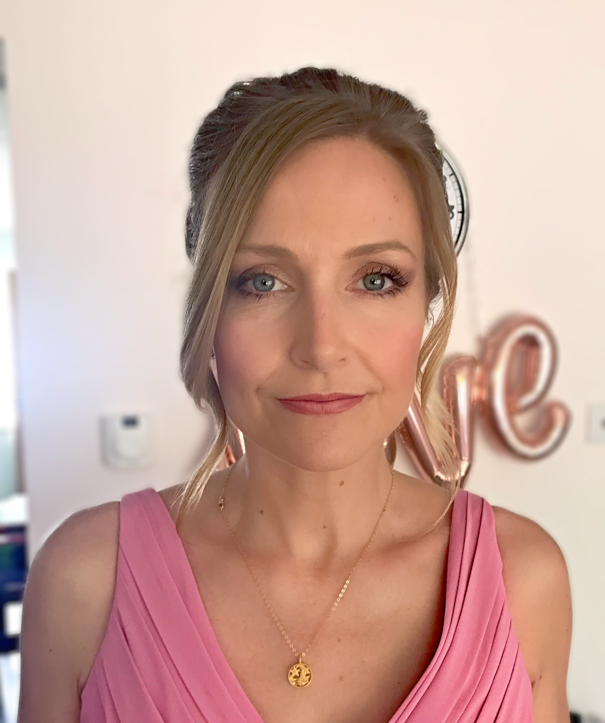 Claire Field Bridal Hair and Make up. A woman with light skin, blonde hair styled in an updo, wearing makeup, gold necklace with a pendant, and pink dress, standing indoors with a blurred background including a metallic "love" sign and a wall clock.