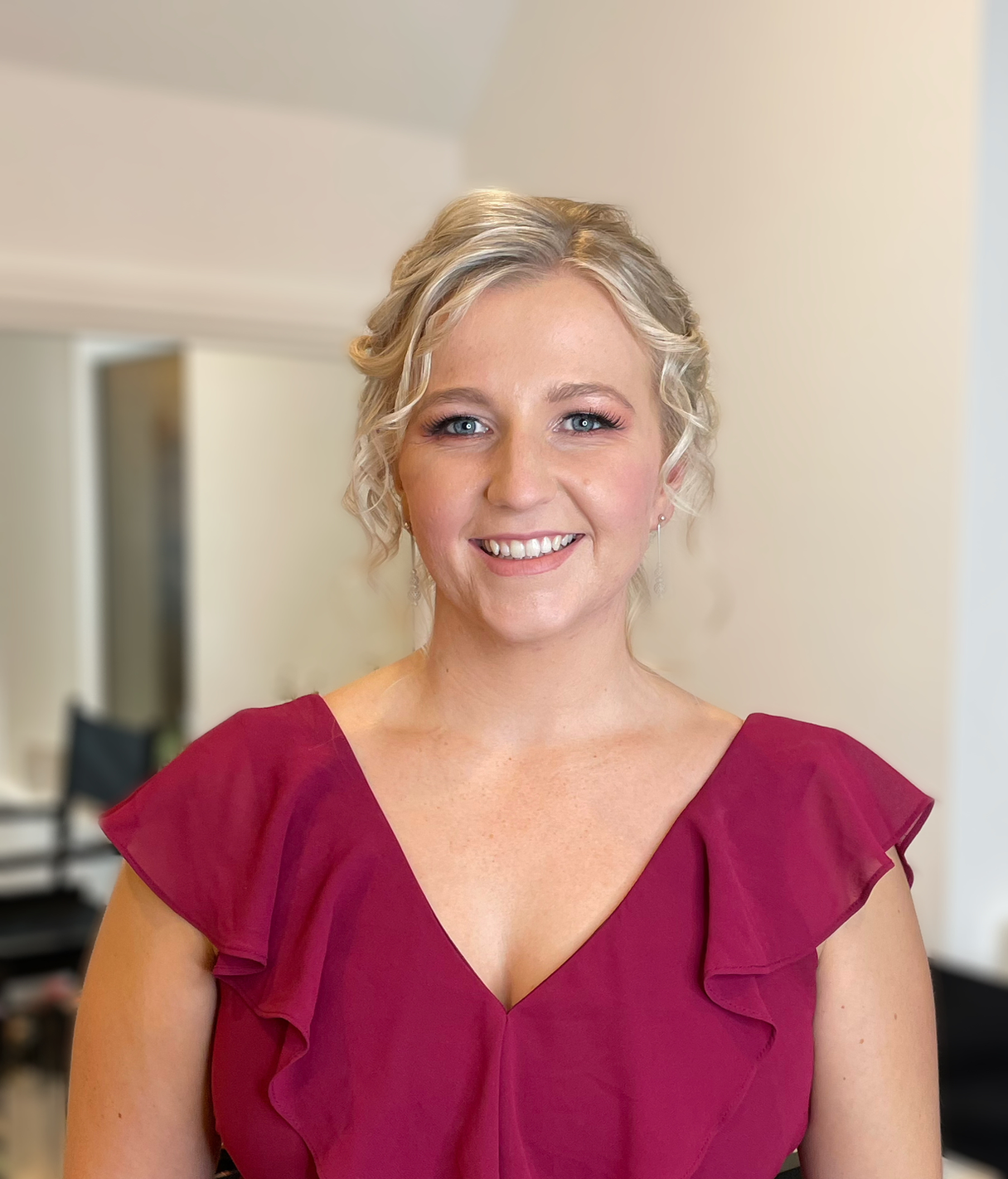 Claire Field Bridal Hair and Make up. A woman with blonde curly hair wearing a magenta dress and earrings, smiling indoors.