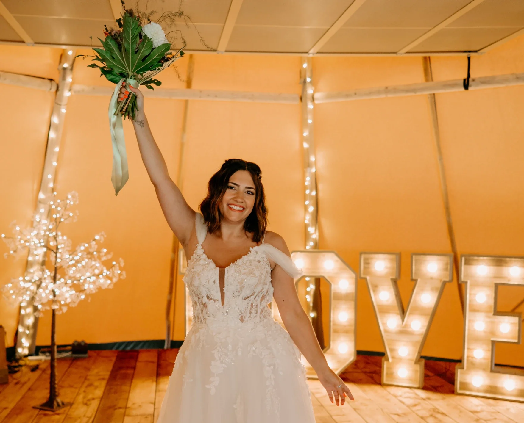 Claire Field Bridal Hair and Make up. A woman in a white wedding dress holding a bouquet of flowers in a decorated indoor venue.