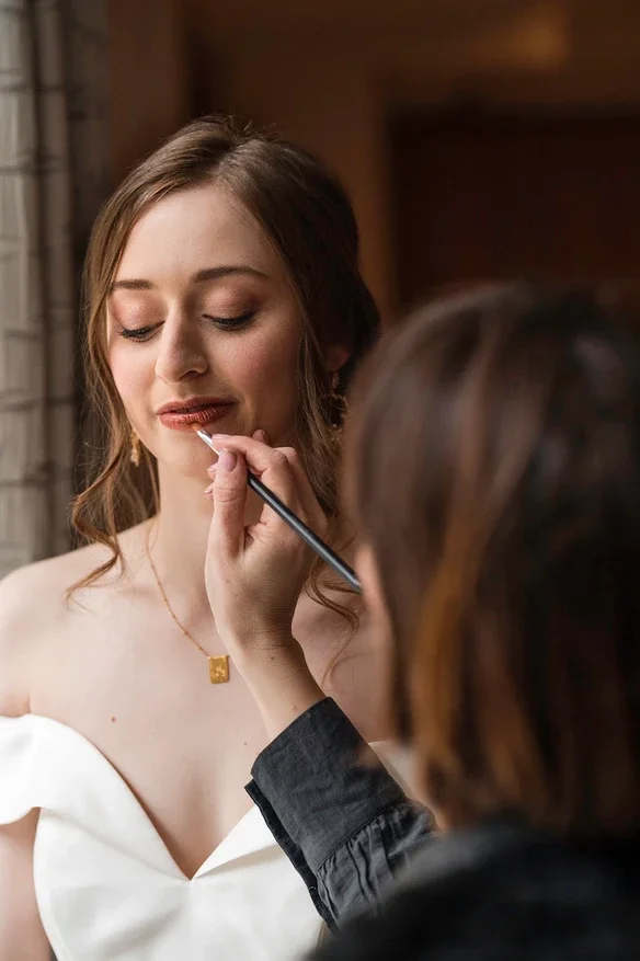 Claire Field Bridal Hair and Make up. A woman with long brown hair getting her makeup done, wearing a strapless white dress and a gold necklace, with her eyes closed and a slight smile.