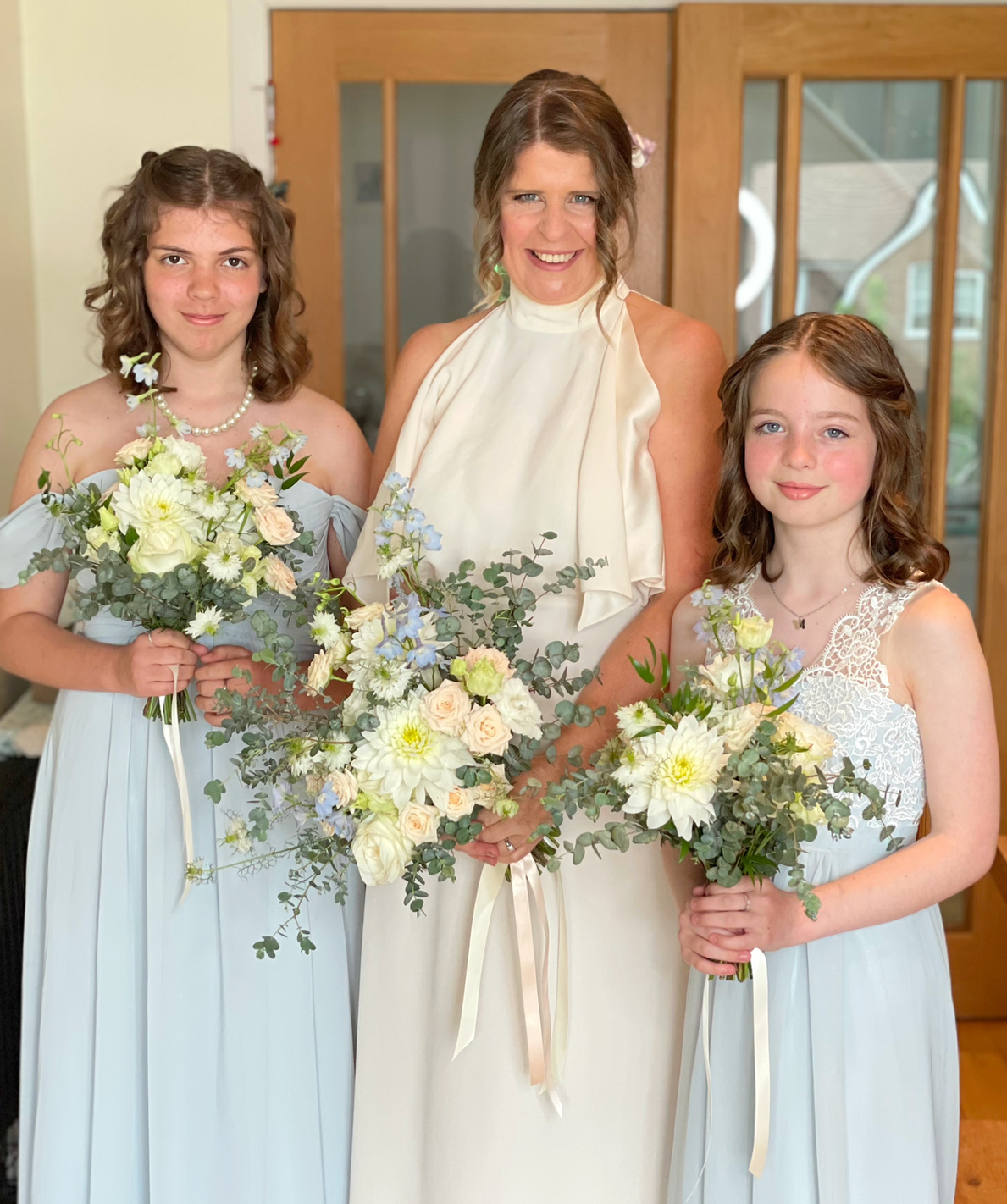 Claire Field Bridal Hair and Make up. A woman and two young girls dressed in white holding bouquets of flowers, standing indoors in front of wooden door with glass panels.