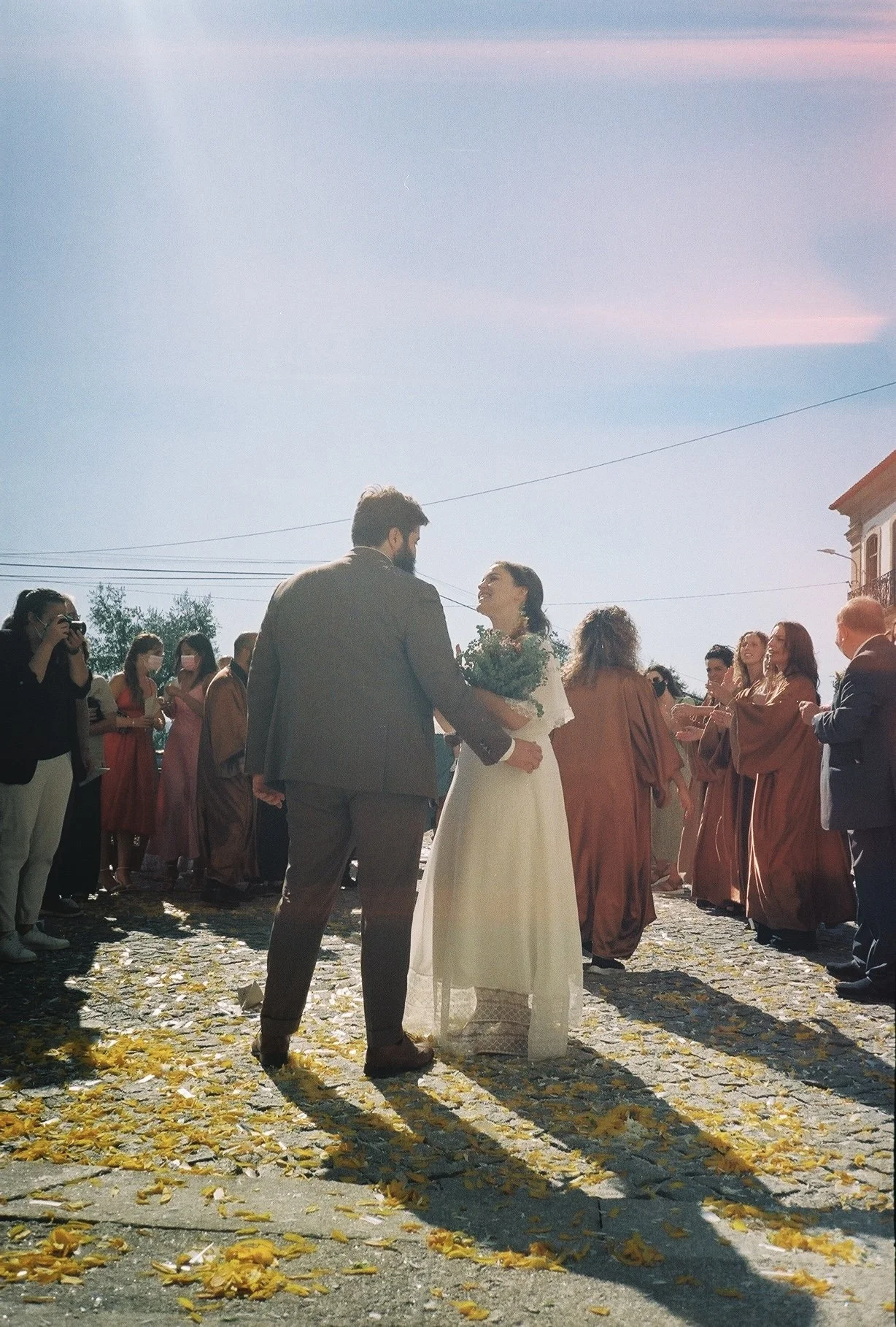 A wedding ceremony outdoors on a cobblestone street with yellow leaves, under a pink and blue sky, featuring a bride in a white dress holding a bouquet and a groom in a gray suit, surrounded by guests.