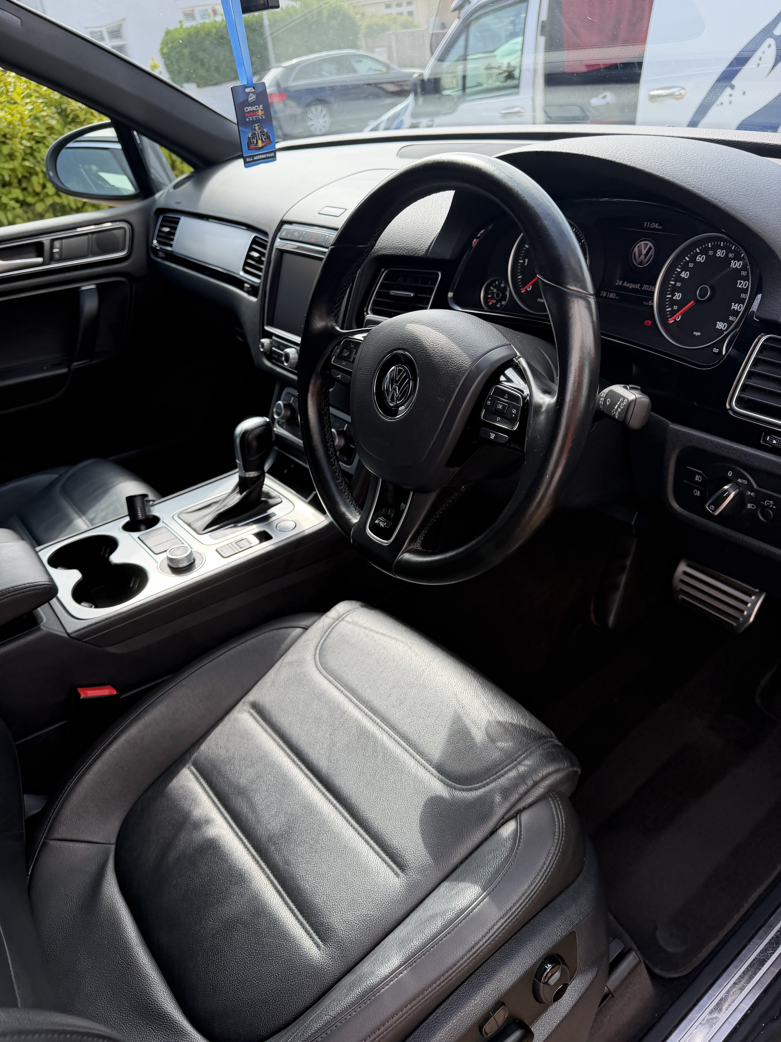 Interior of a Volkswagen vehicle showing the steering wheel, dashboard, center console, black leather seat, and a gear shift lever. Outside the windshield, cars and greenery are visible.
