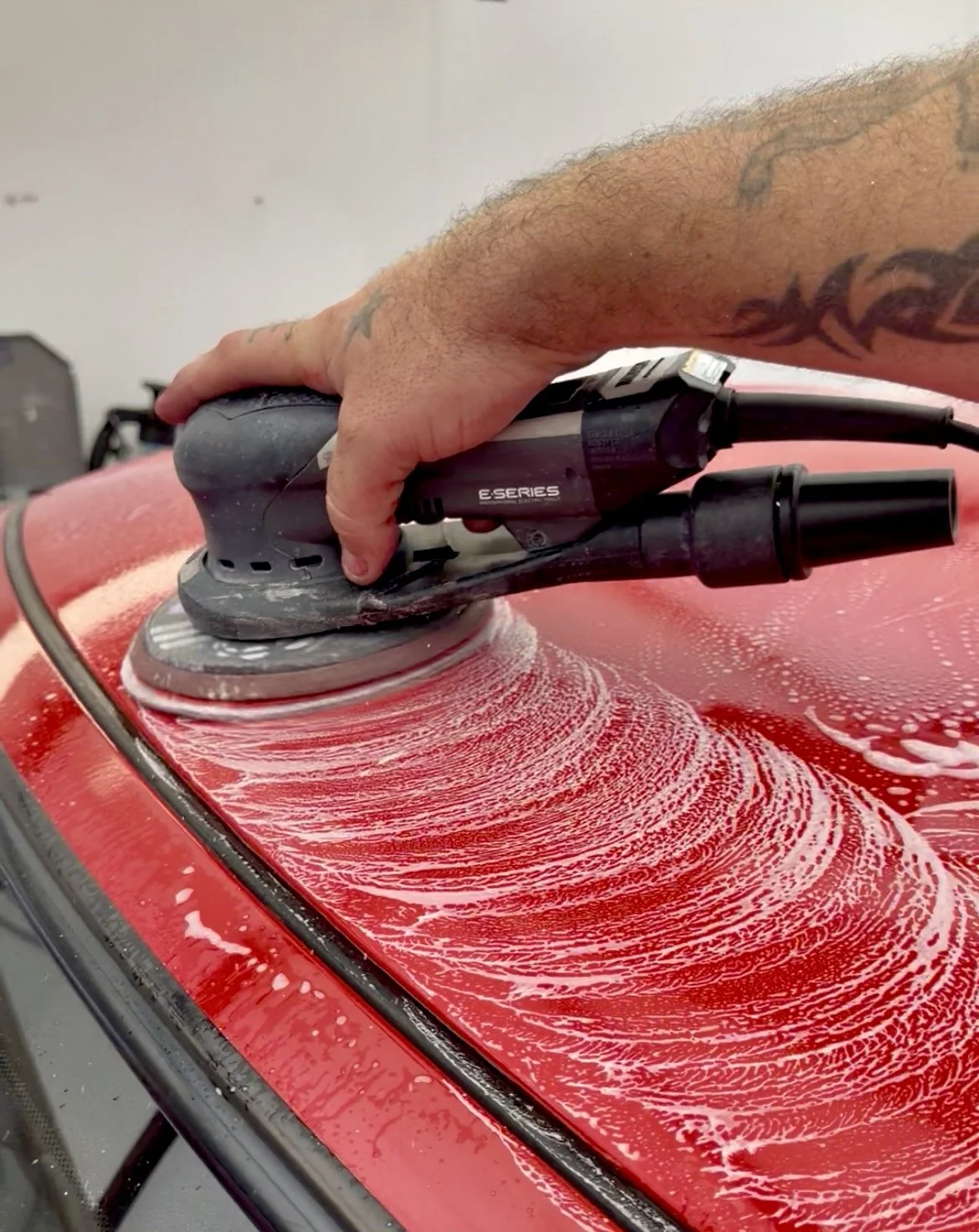 A person is polishing a red car with an electric buffer tool, creating soap suds and foam on the car's surface.