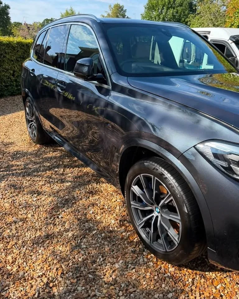 A black BMW SUV parked on a gravel surface with trees and a clear blue sky in the background.