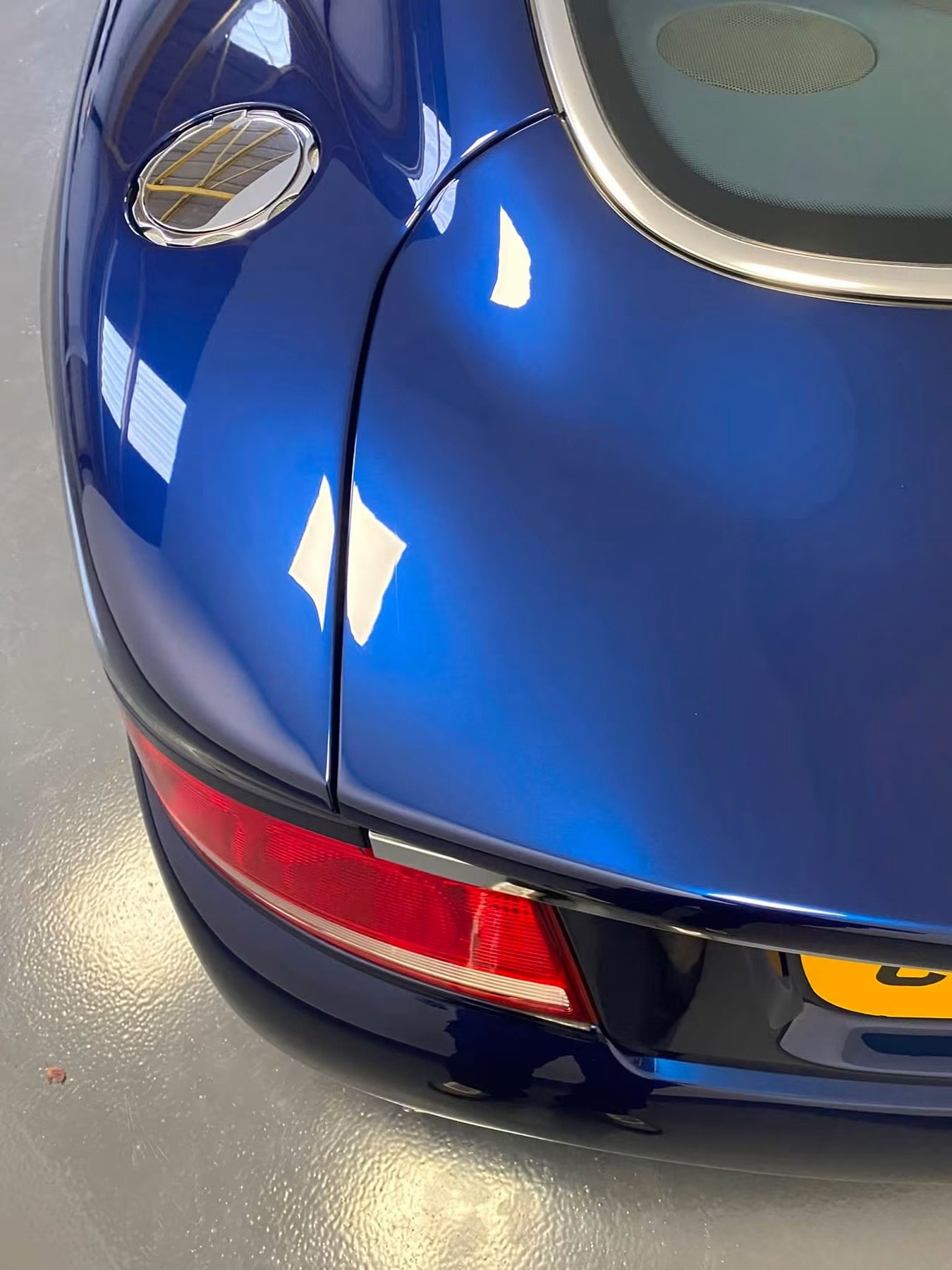 Close-up of the rear end of a blue car with a shiny gas cap cover, a red taillight, and a license plate, parked on a gray concrete floor.