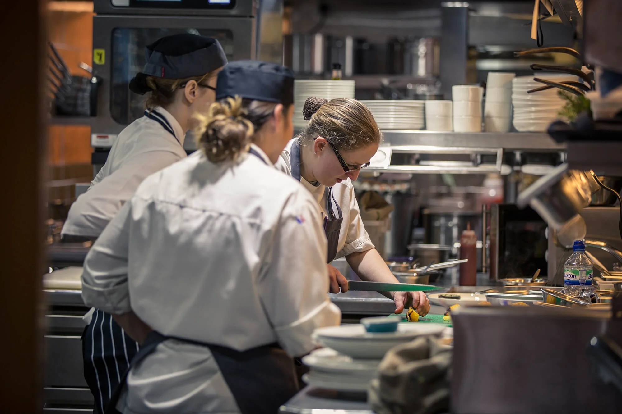 Group of chefs or cooks working in a professional kitchen, preparing food at a counter with stacks of plates in the background.