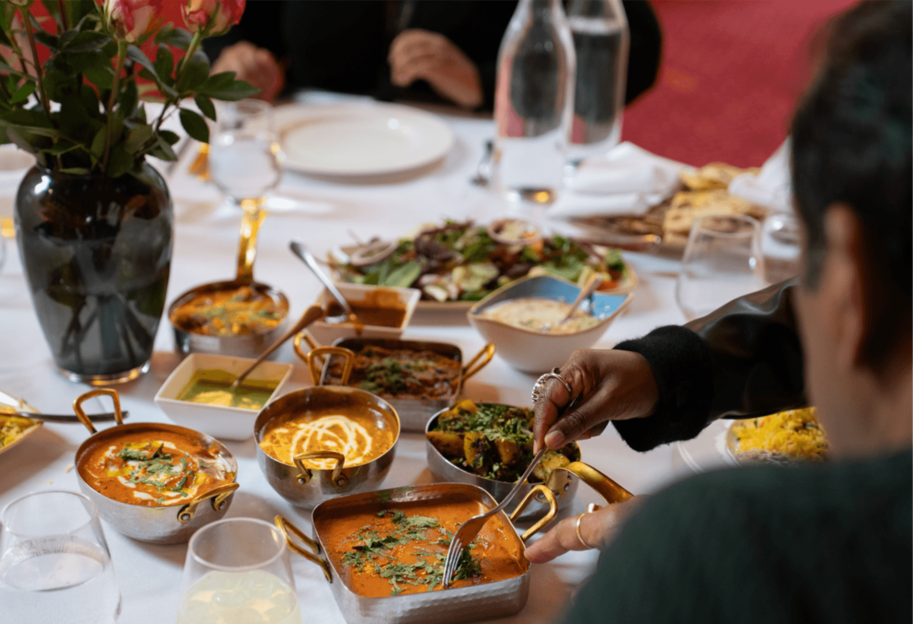 A dining table with various Indian dishes and bowls of curries, a person serving food, a potted plant, and glasses of water. The table is set for a meal with plates and utensils.