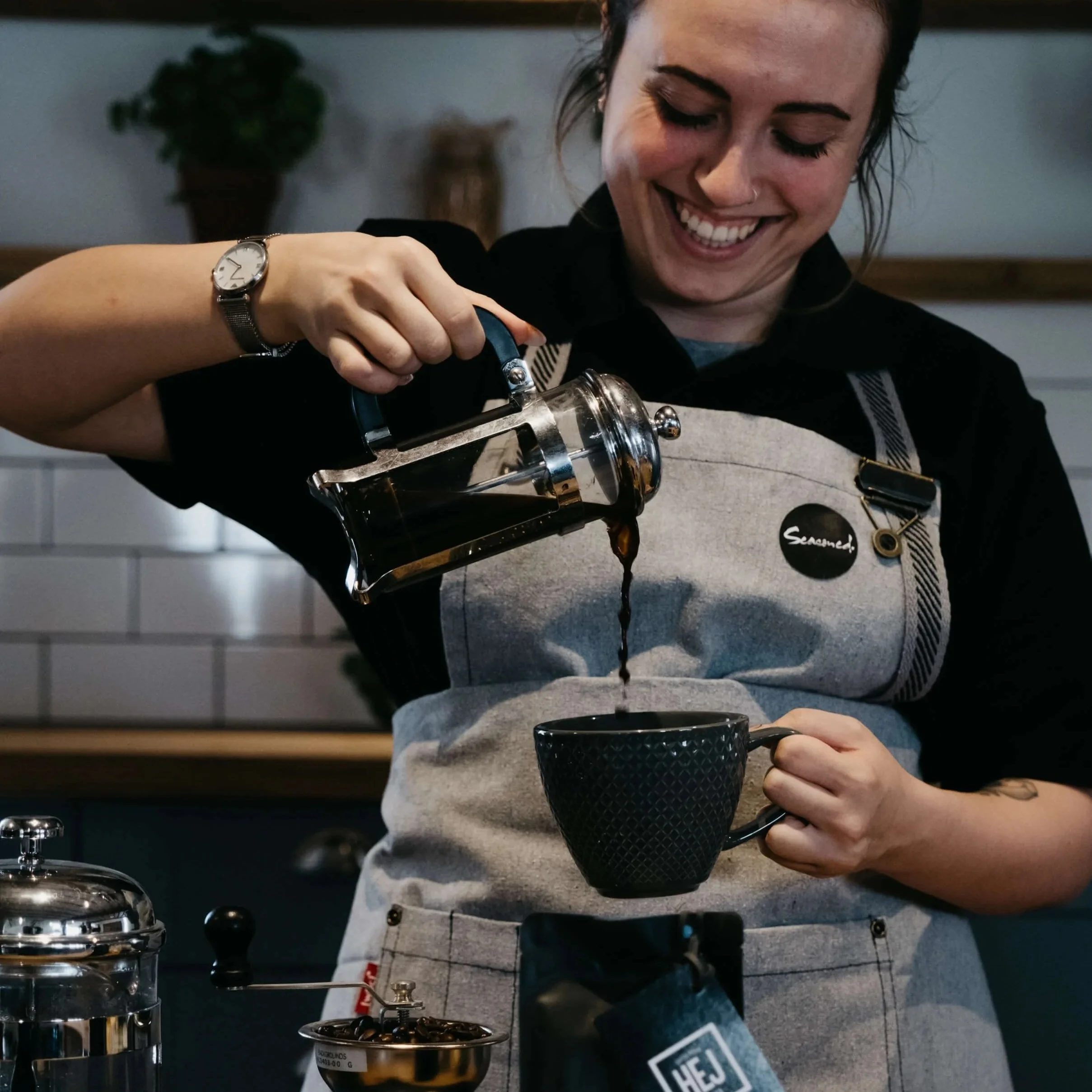 A woman smiling while holding a coffee tamper above a portafilter in a coffee shop or barista setting. She is wearing a watch on her left wrist and an apron labeled 'SEARCH'.