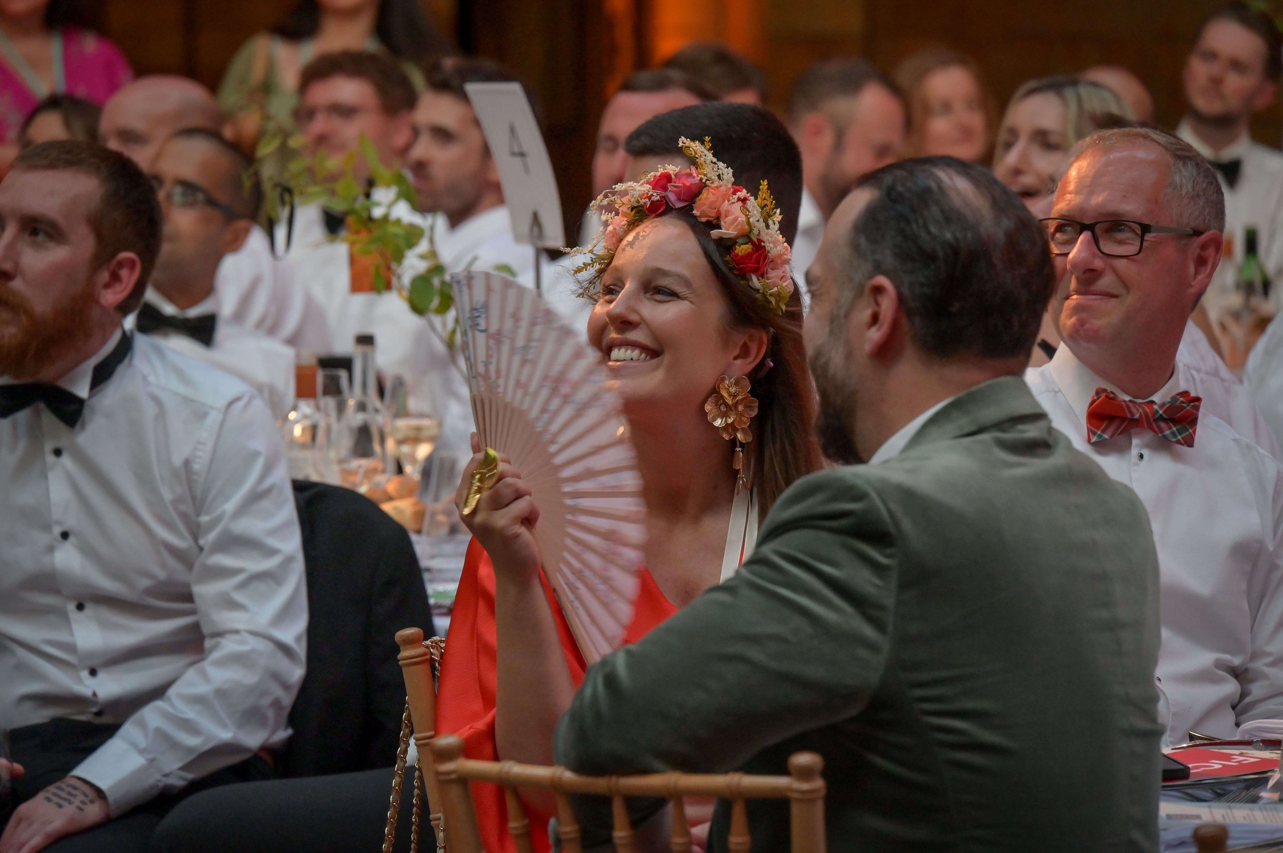 Celebrants at a wedding reception, including a woman in a floral crown and coral dress smiling, holding a fan.