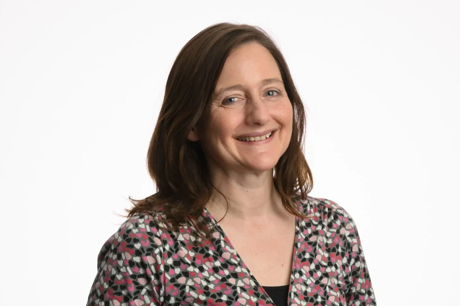 A smiling woman with shoulder-length brown hair, wearing a patterned blouse with pink, black, and white hearts, against a plain white background.