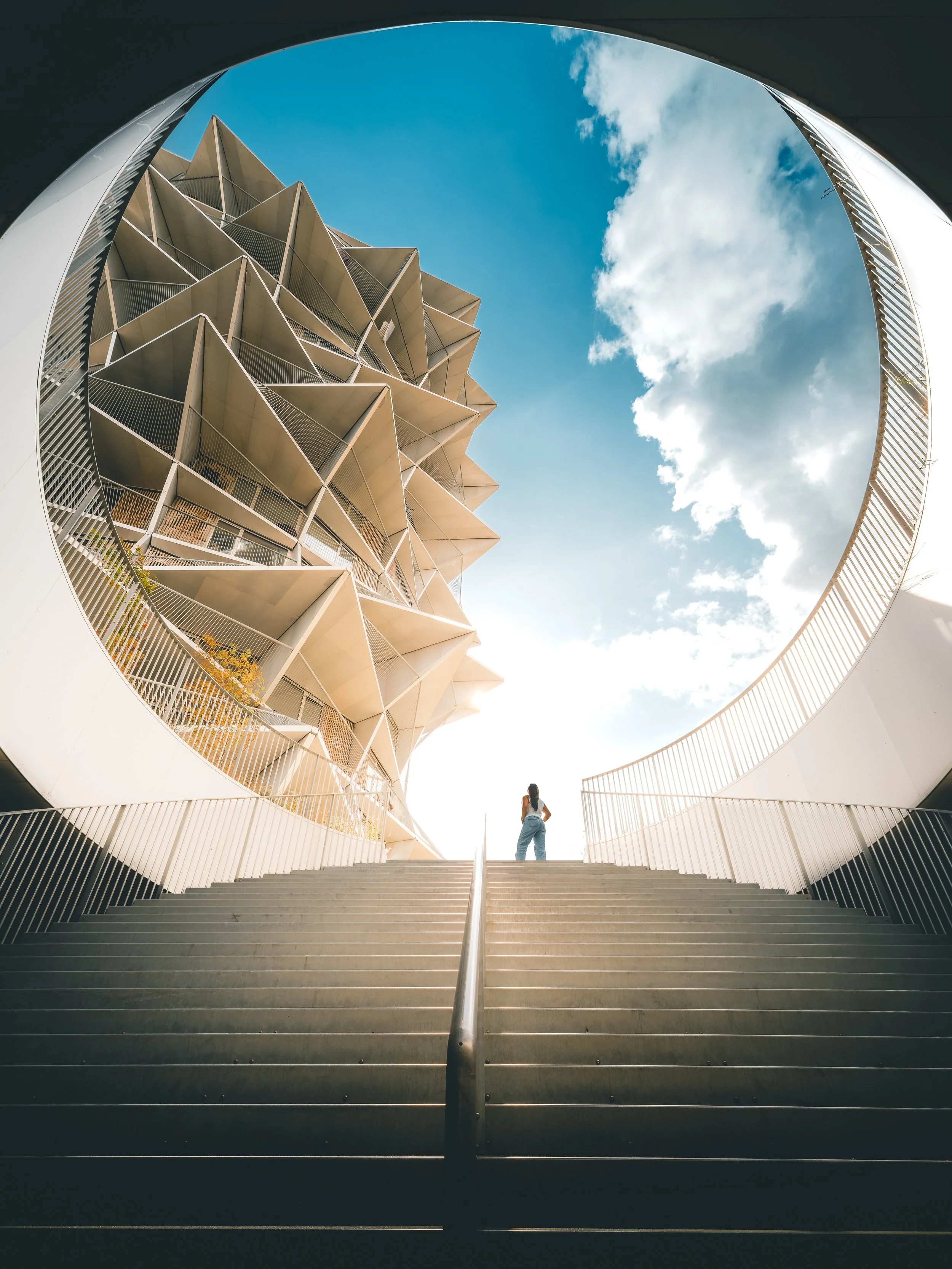 De afbeelding toont een personage die op de bovenverdieping van een moderne architectuur staat, gezien vanaf de onderkant van een brede trap. De trap leidt omhoog onder een ronde opening in het plafond, waar een uniek, kubusvormig gebouw met meerdere