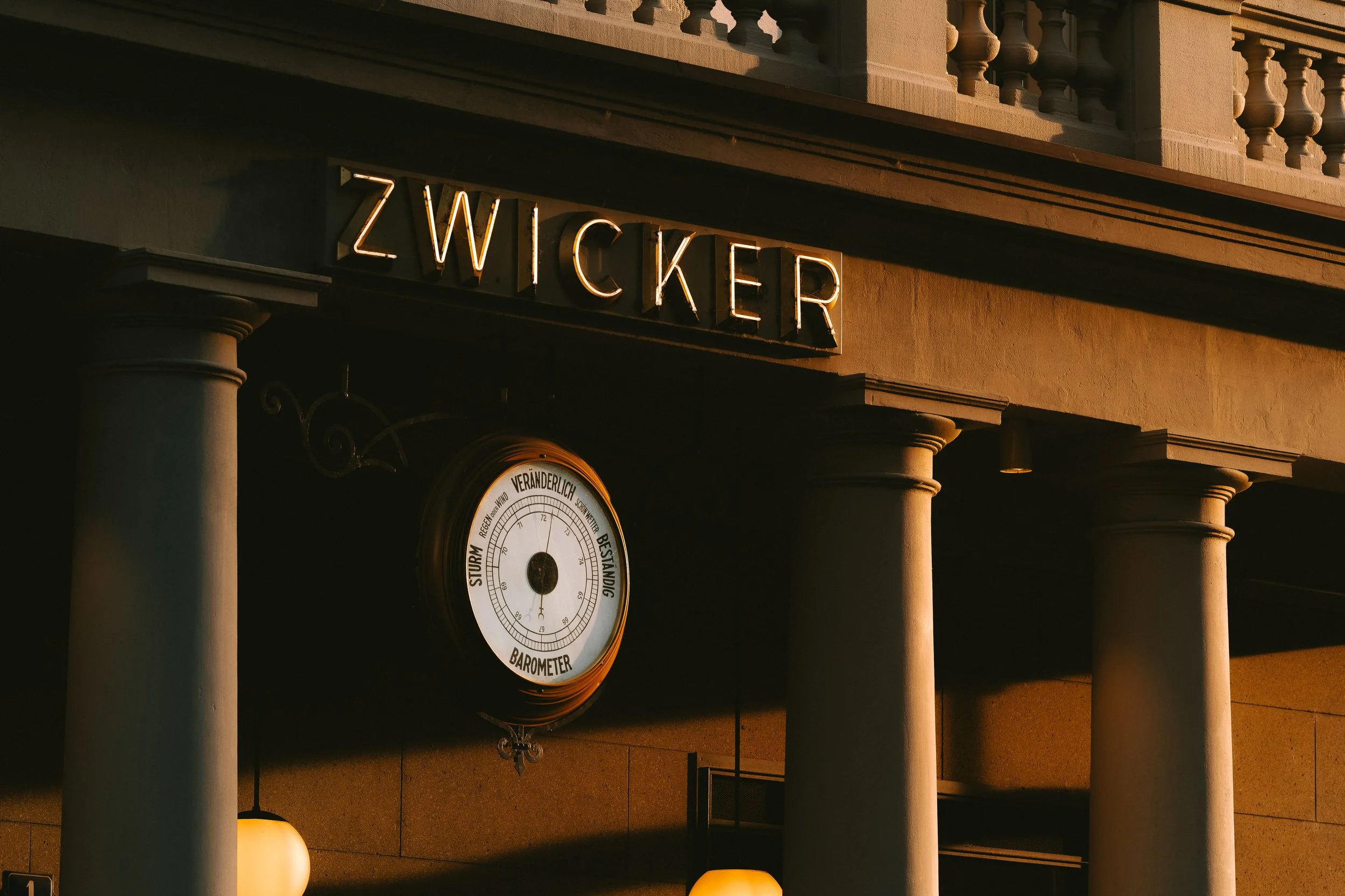 Uitzicht van een gebouw met neonletters die 'Zwick' spellen, verlicht door de zon, met een barometer onderaan en zandkleurige muren en kolommen.