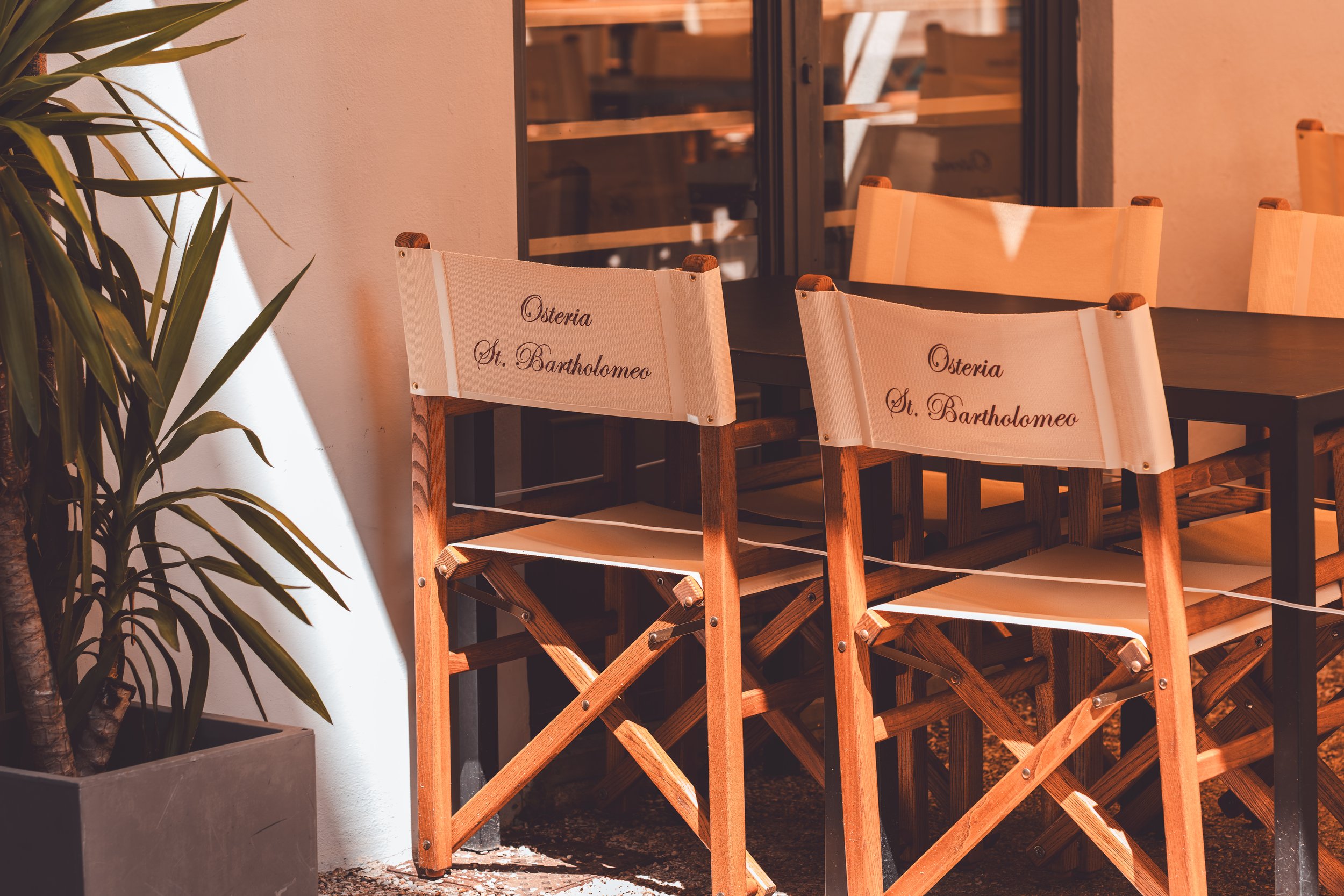 Twee houten stoelen met tekst 'Osteria St. Bartholomeo' in een restaurant met planten en een glaswand op de achtergrond.