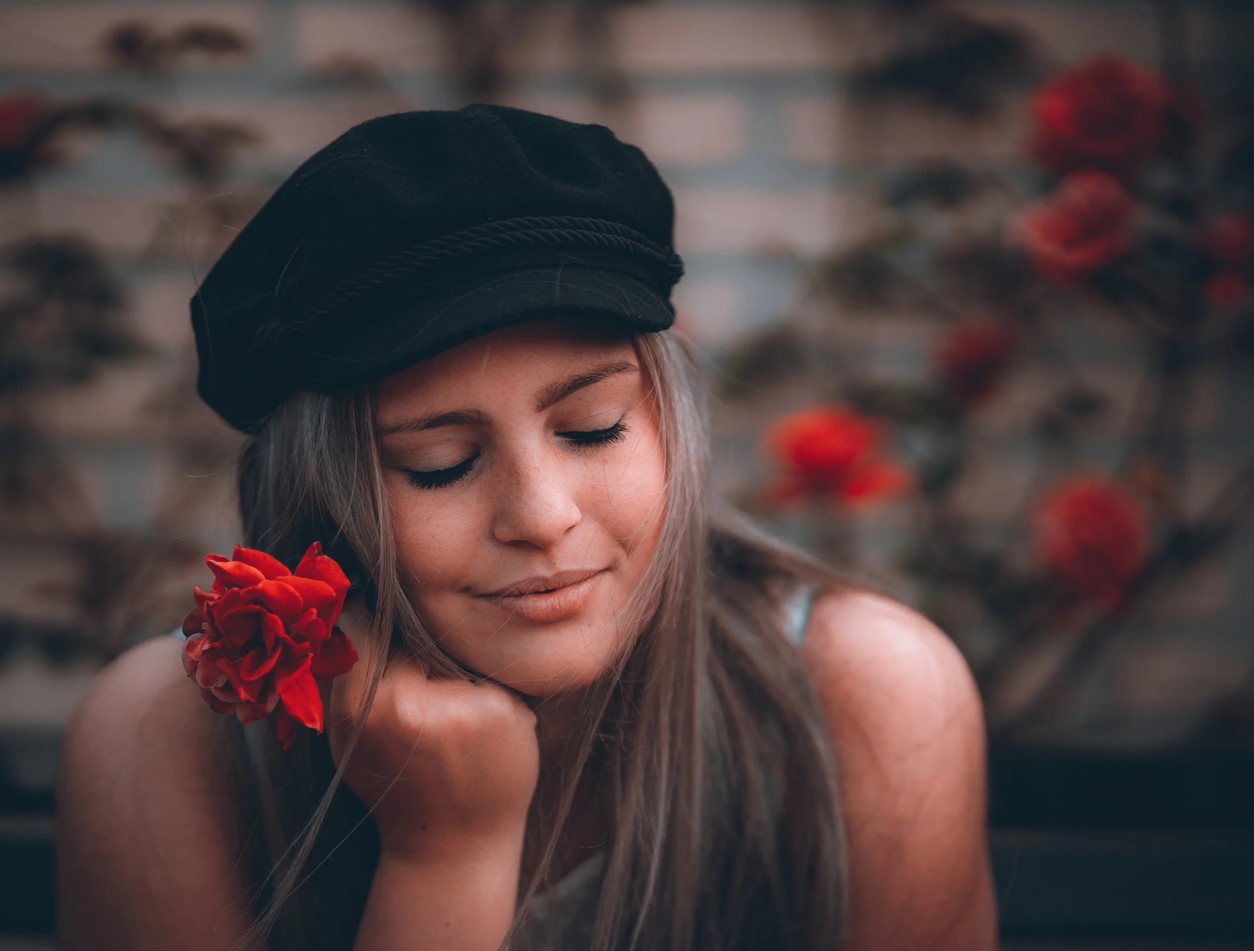 Close-up van een vrouw met gesloten ogen, een zwarte hoed en een rode bloem in haar haar, in een natuurlijke buitenomgeving.