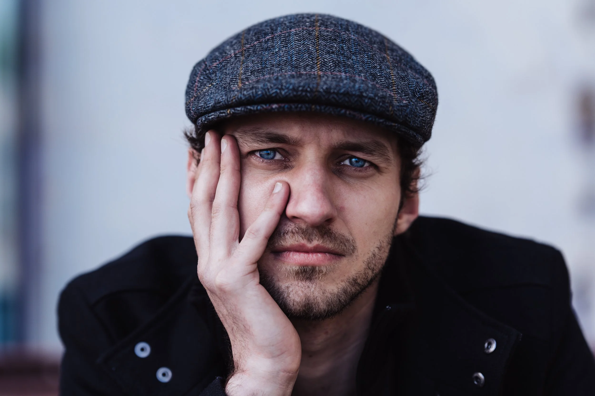 Een man met blauwe ogen draagt een wollige pet en ondersteunt zijn gezicht met zijn hand, terwijl hij naar de camera kijkt.