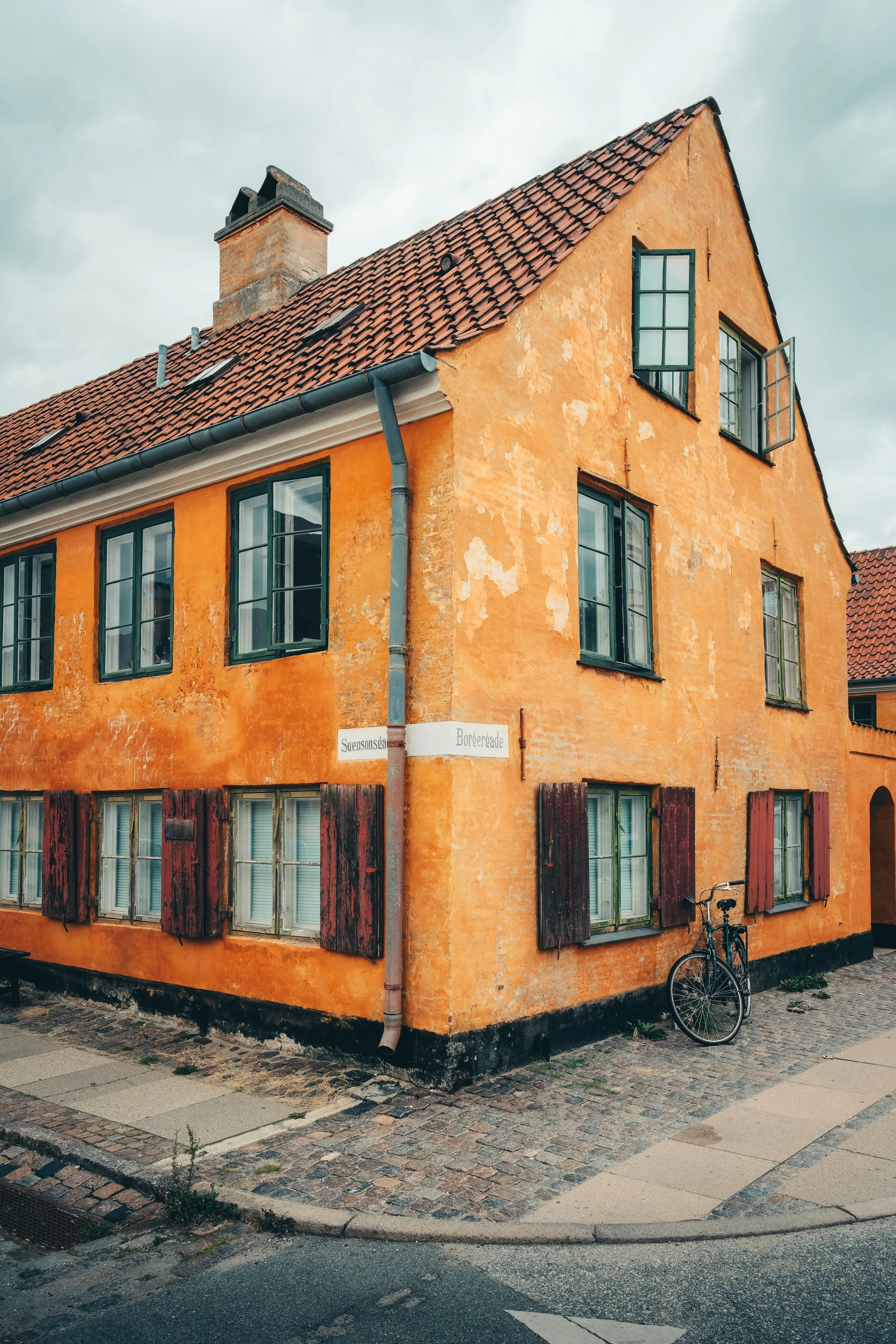 Een oranje gekleurde oude woning met groen geschilderde ramen en houten luiken, en een schoorsteen op het dak. Een fiets staat tegen de muur en er ligt een straatnaambordje met de tekst 'Suensonsga' en 'Borgergave'.