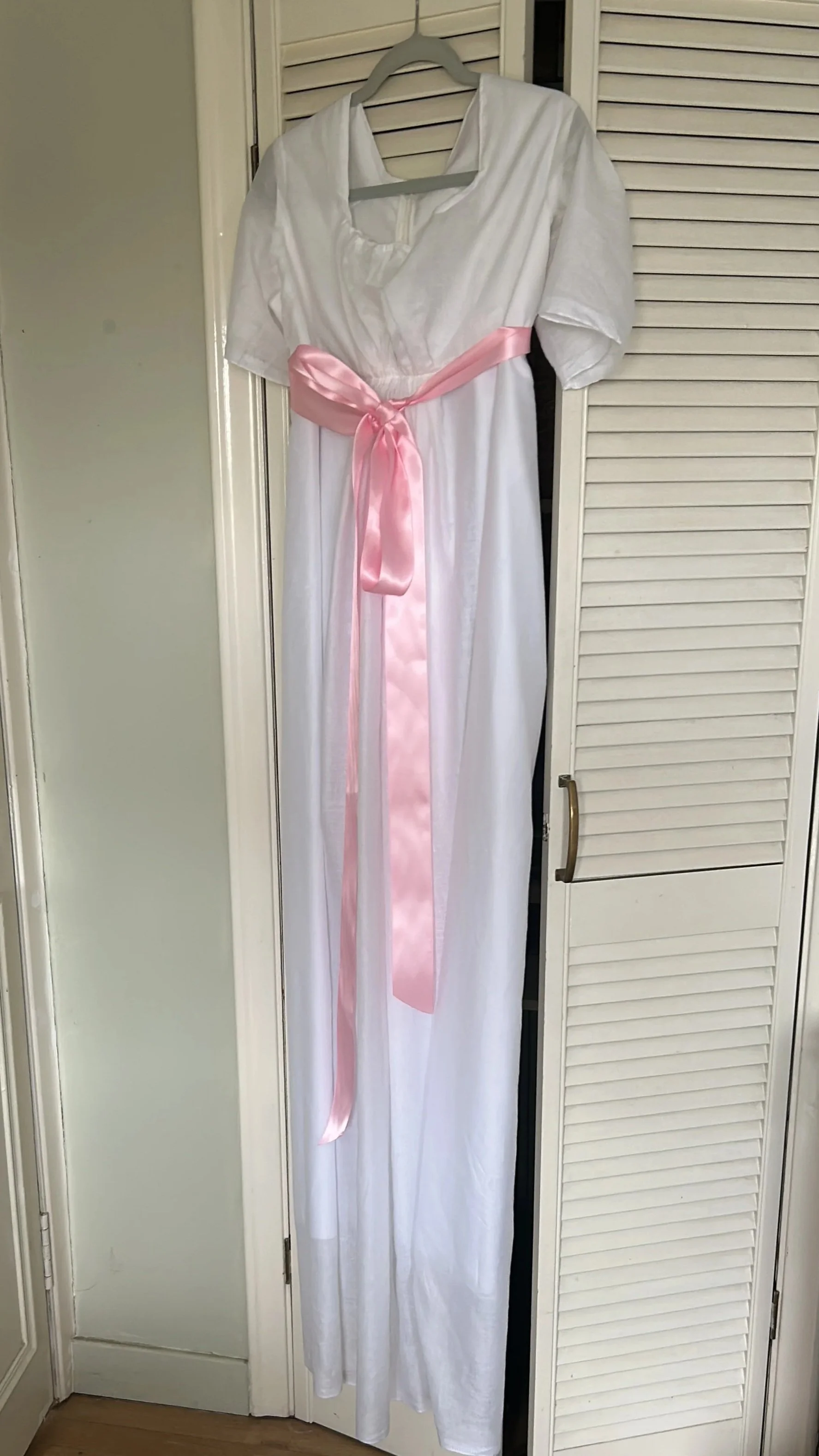 White dress with short sleeves and pink satin sash hanging on a closet door.