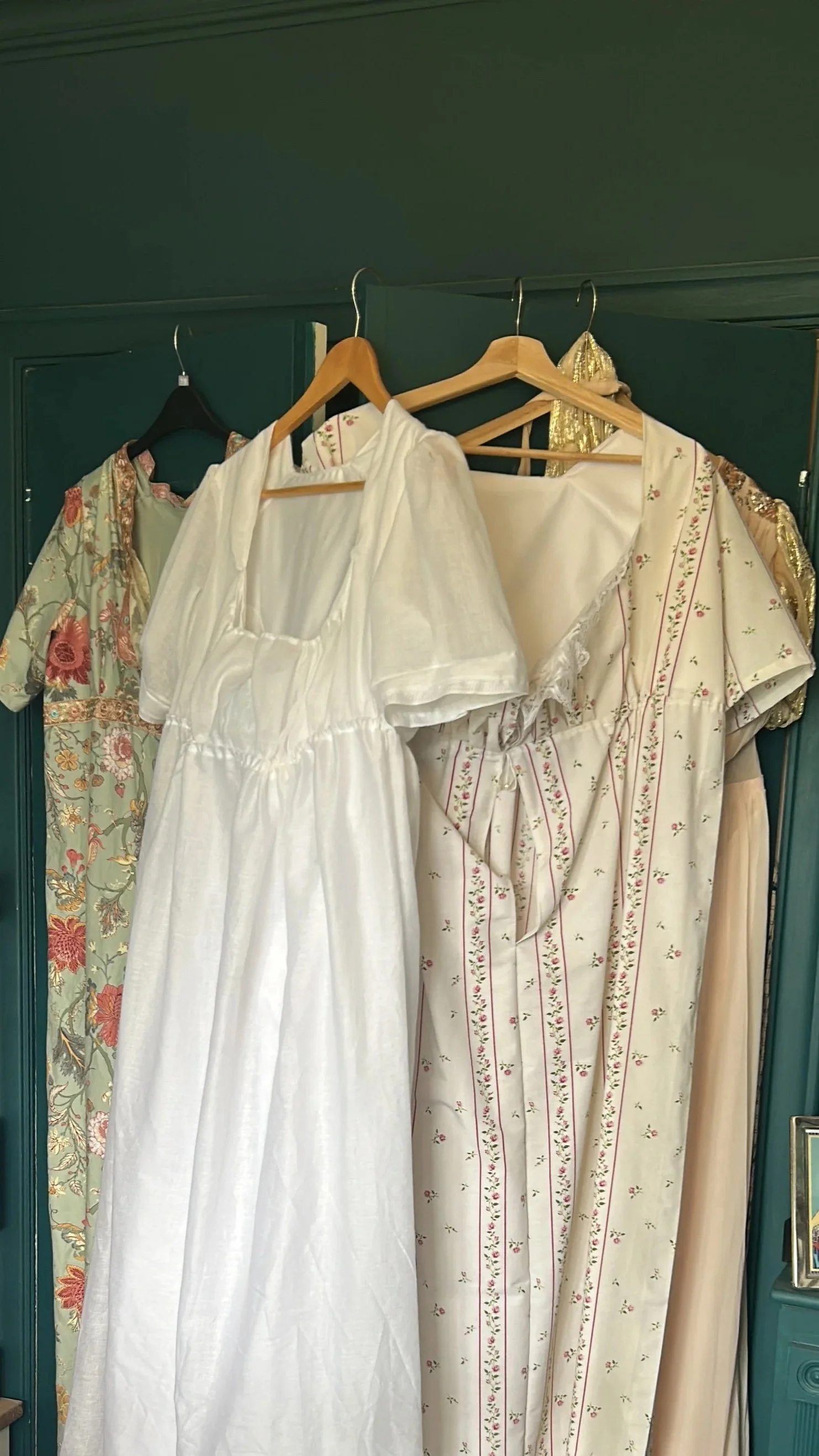 Various dresses hanging on a dark green closet door, including a white dress, a floral dress, and a striped dress with small floral patterns.