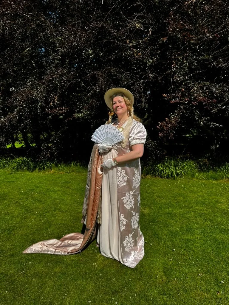 Woman dressed in vintage-style dress with floral patterns, holding a fan, wearing a wide-brimmed hat, standing on a grassy lawn with trees in the background.
