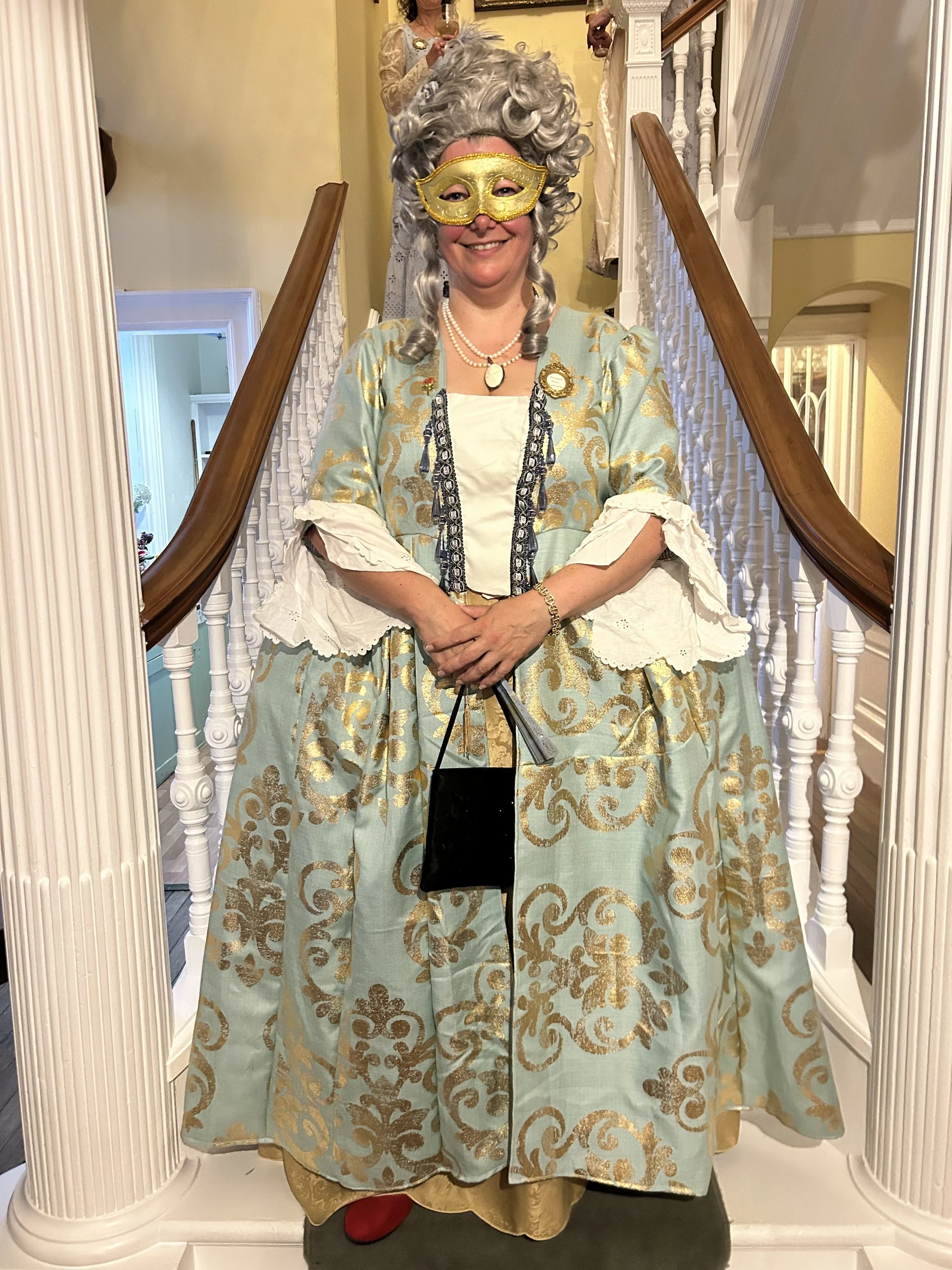 Person dressed in an 18th-century costume with a large, ornate gown, curly silver wig, and a gold mask, standing in front of a staircase with white railings.