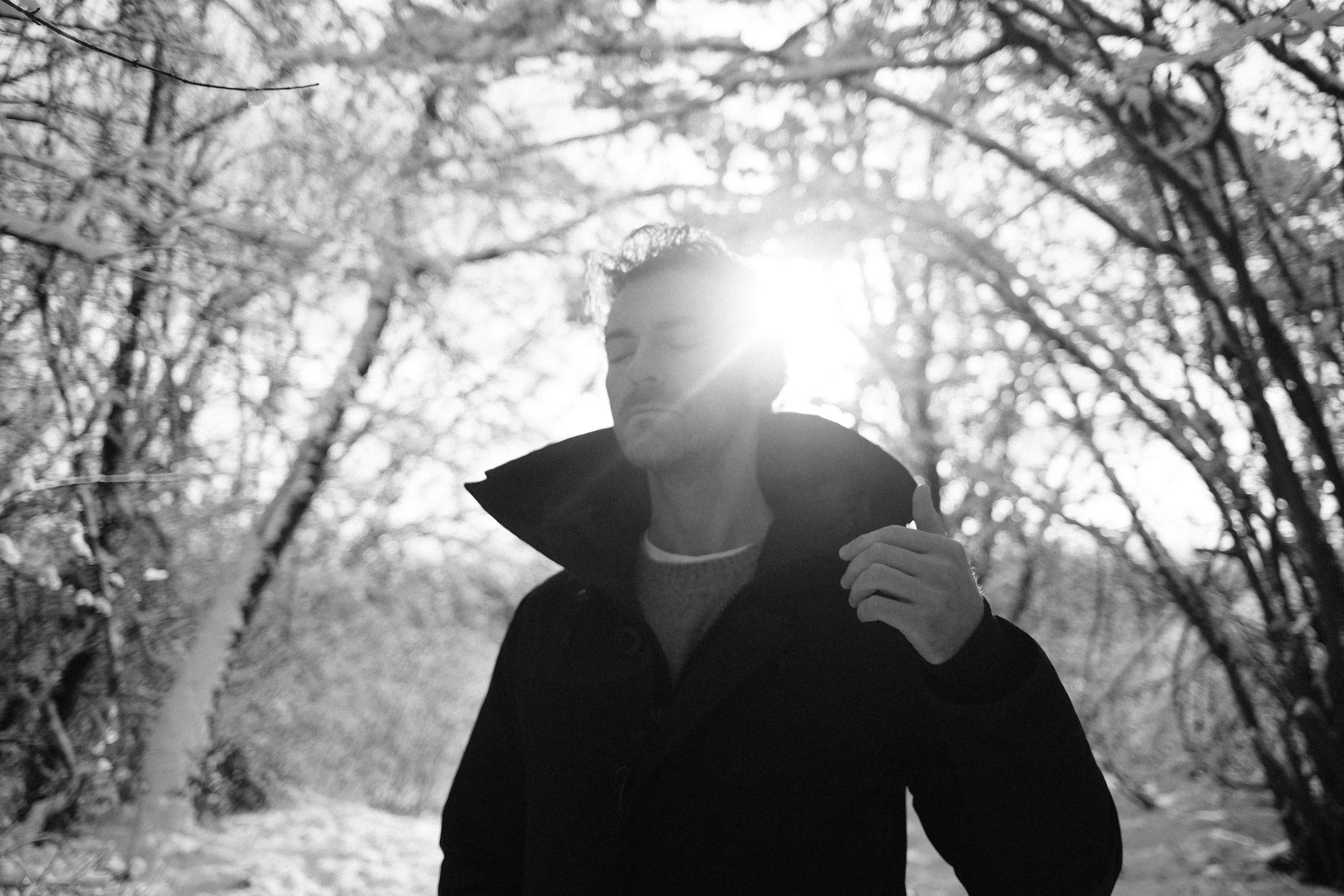 Schwarz-Weiß-Foto eines Mannes im Freien, der eine Jacke trägt und durch eine Baumallee geht, Sonnenlicht im Hintergrund.