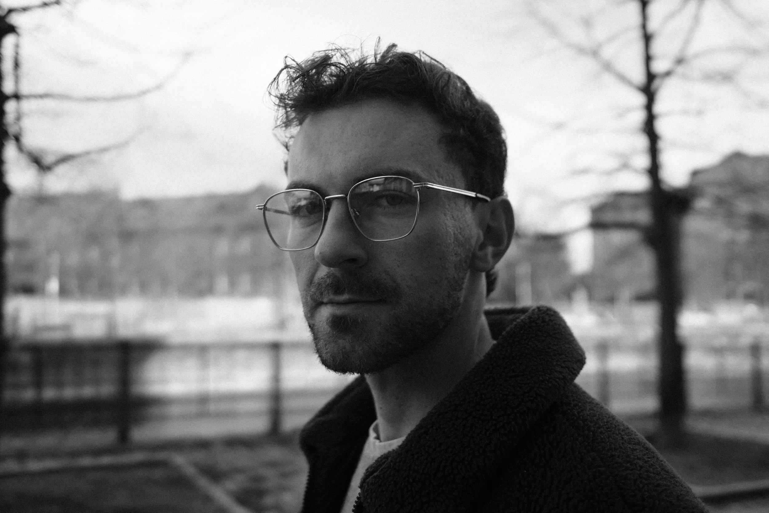 Schwarz-weiß-Foto eines Mannes mit Brille, der nach rechts blickt, im Hintergrund sind Bäume und ein Zaun sichtbar.