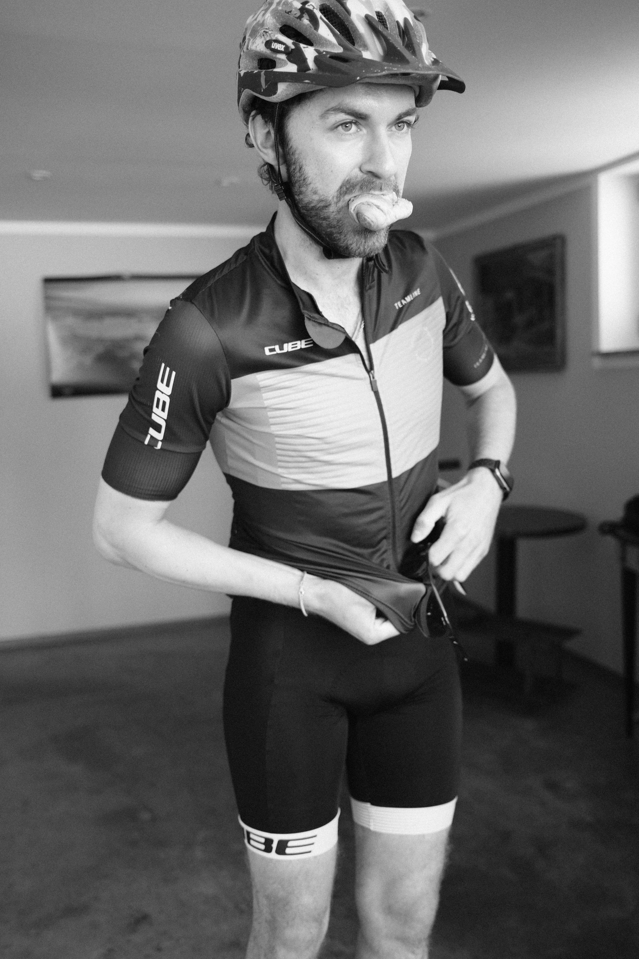 Mann in Fahrradhelm, Fahrradtrikot, Shorts und Kompressionsstrümpfen im Fitnessstudio, isst einen Snack.