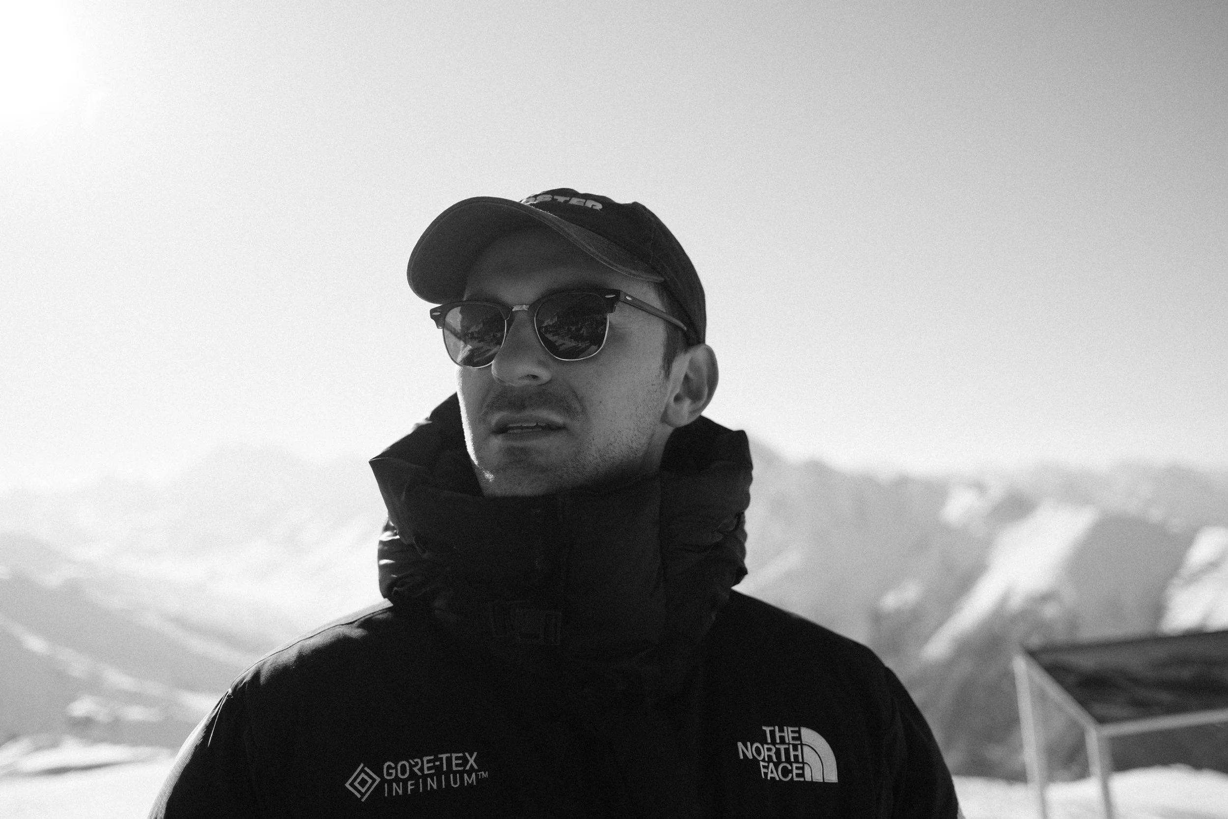 Ein Mann trägt eine schwarze Jacke mit Logo von The North Face und Gore-Tex, eine schwarze Kappe, Sonnenbrille und steht in einer verschneiten Berglandschaft.