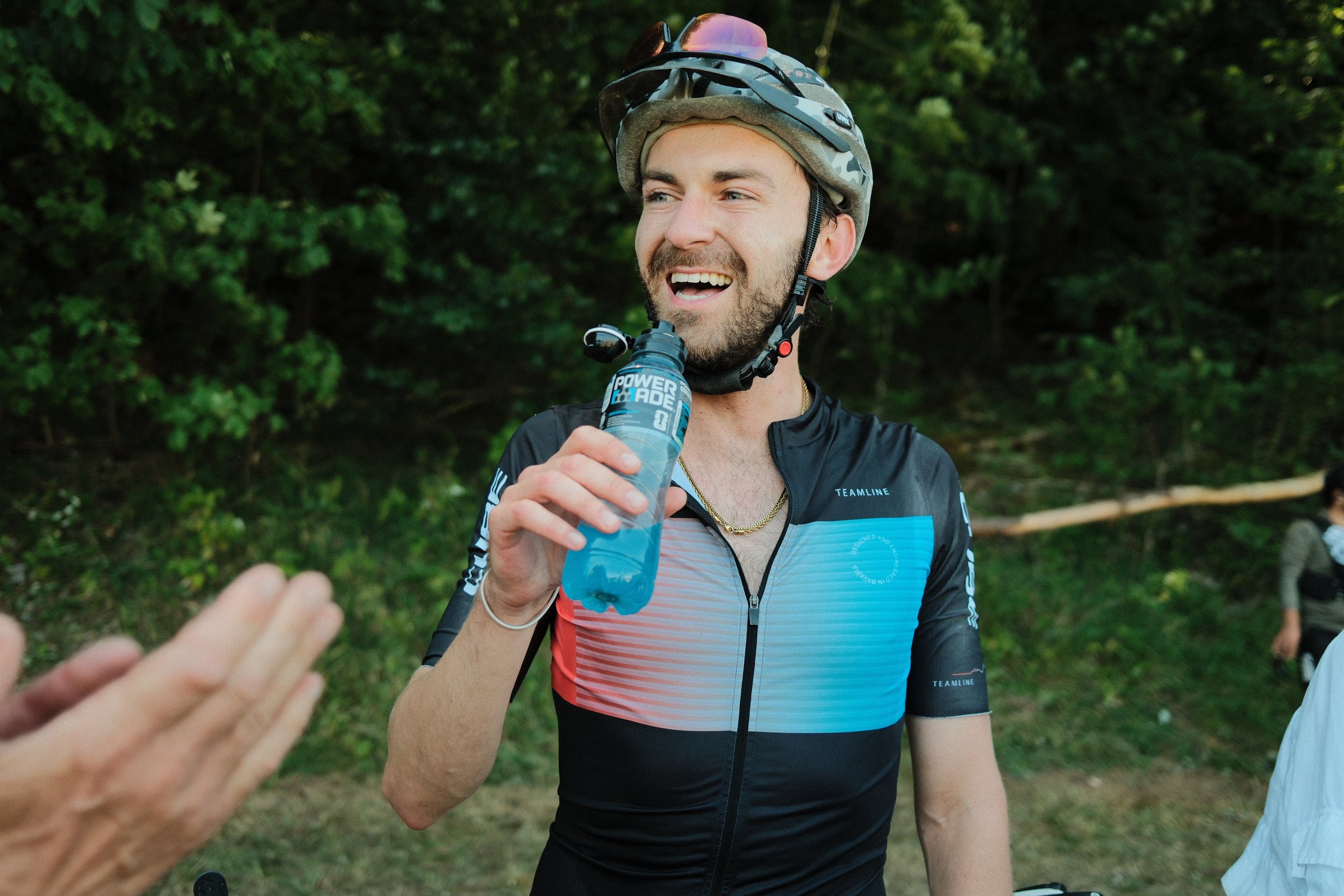 Ein lächelnder junger Mann trägt einen Fahrradhelm und ein Fahrradtrikot, während er in einer grünen, bewaldeten Umgebung Wasser trinkt.
