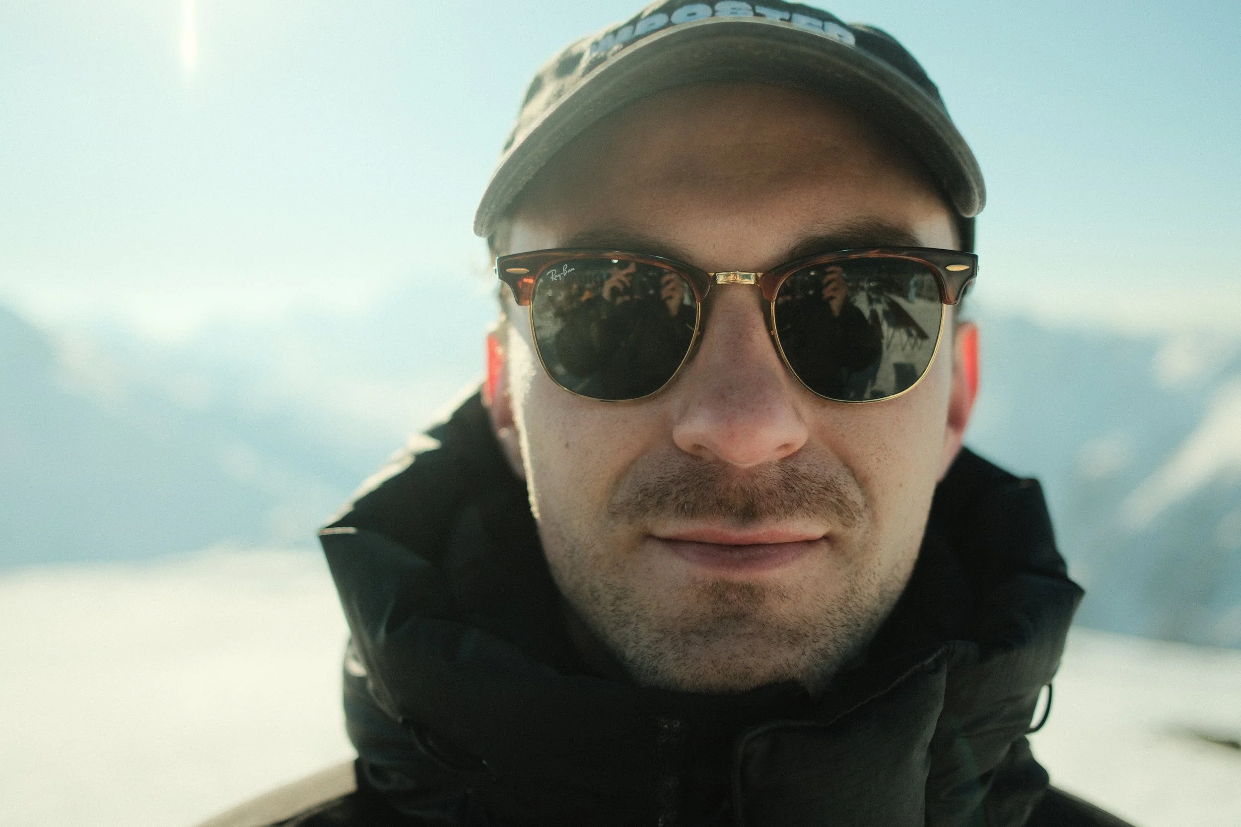 Nahaufnahme eines Mannes mit Sonnenbrille, Kappe und dunkler Jacke, im Schnee bei bewölktem Himmel.
