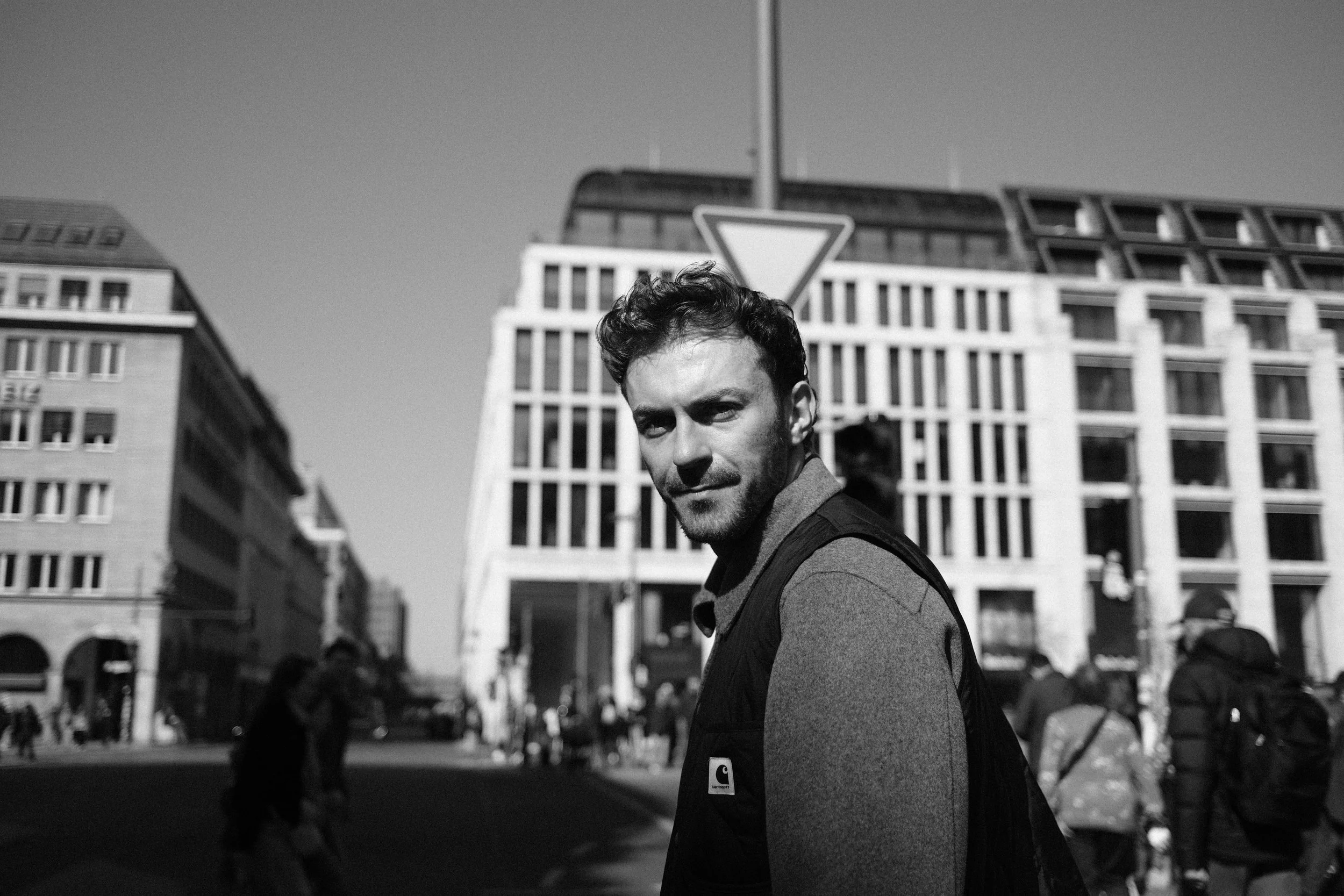 Schwarz-Weiß-Foto eines Mannes mit dunklem Haar und Bart, der in einer Stadt steht, umgeben von Hochhaushäusern und mit Menschen im Hintergrund.