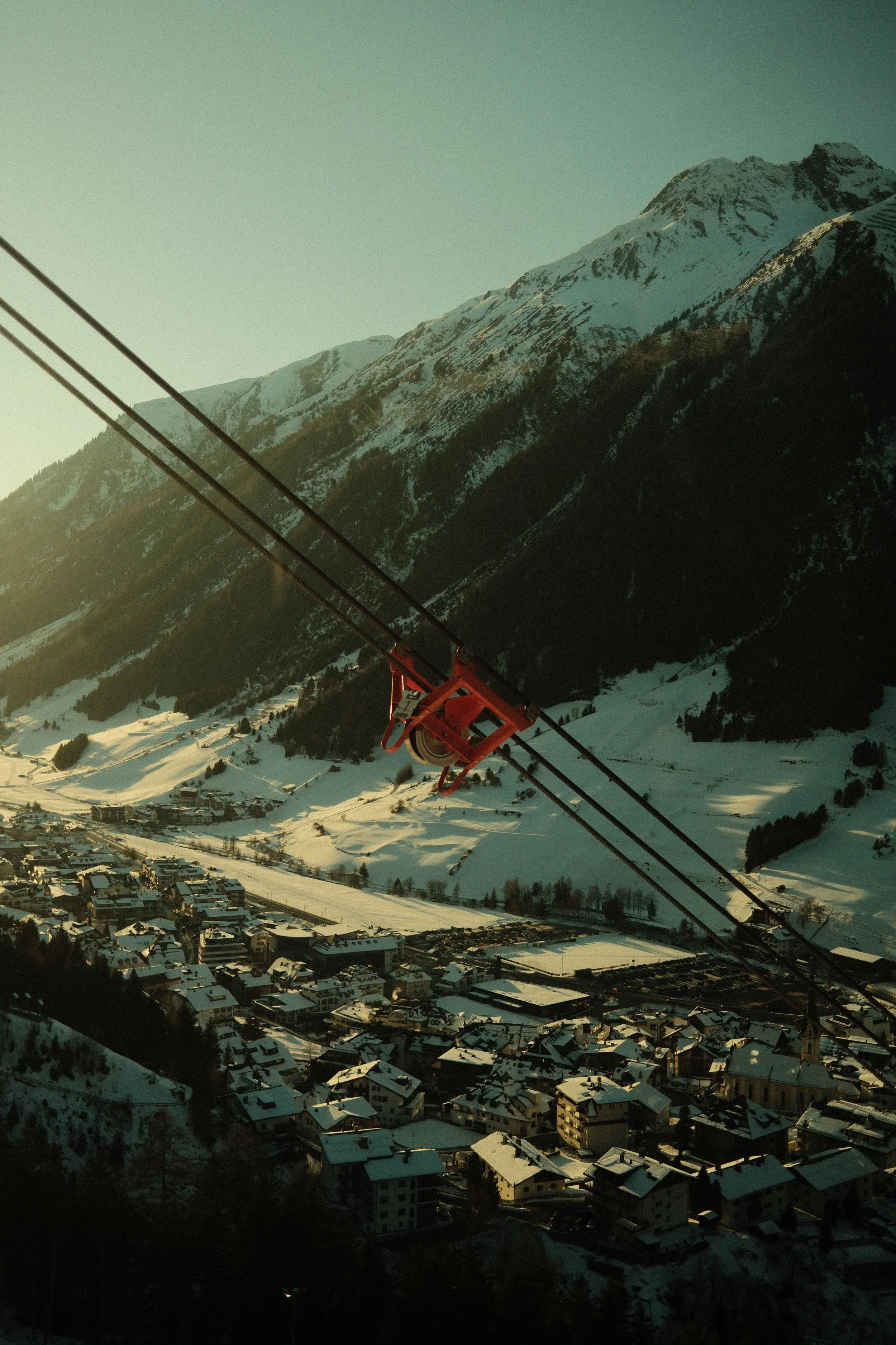 Blick auf eine verschneite Stadt am Fuße eines Berges, mit einem roten Lift und Seilen im Vordergrund, bei Sonnenuntergang