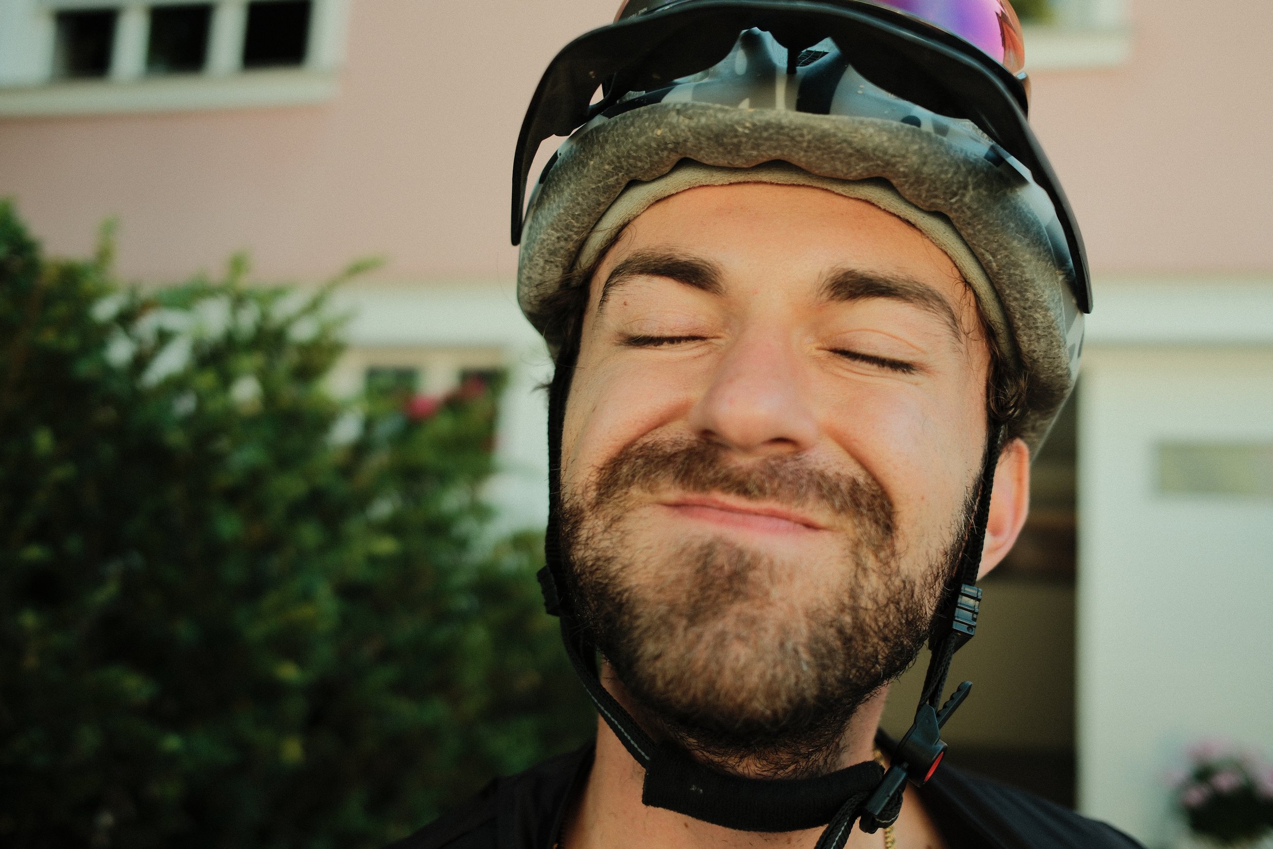 Nahaufnahme eines lachenden Mannes mit Fahrradhelm, geschlossene Augen, dunkle Haare und Bart, im Hintergrund ein Haus und einige grüne Pflanzen.