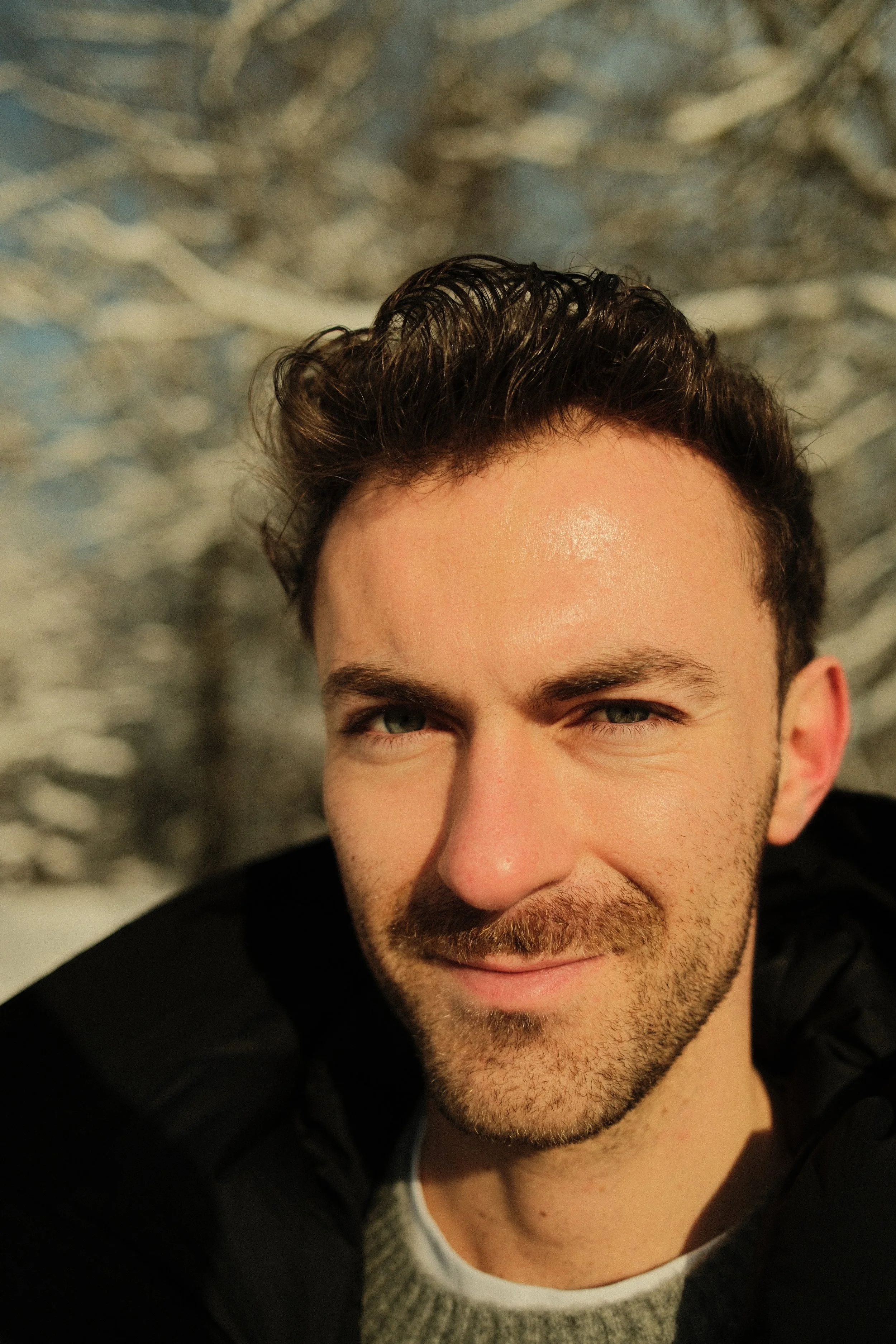 Nahaufnahme eines Mannes mit dunklen, gewellten Haaren, einem Bart und blauen Augen, der lächelt.
