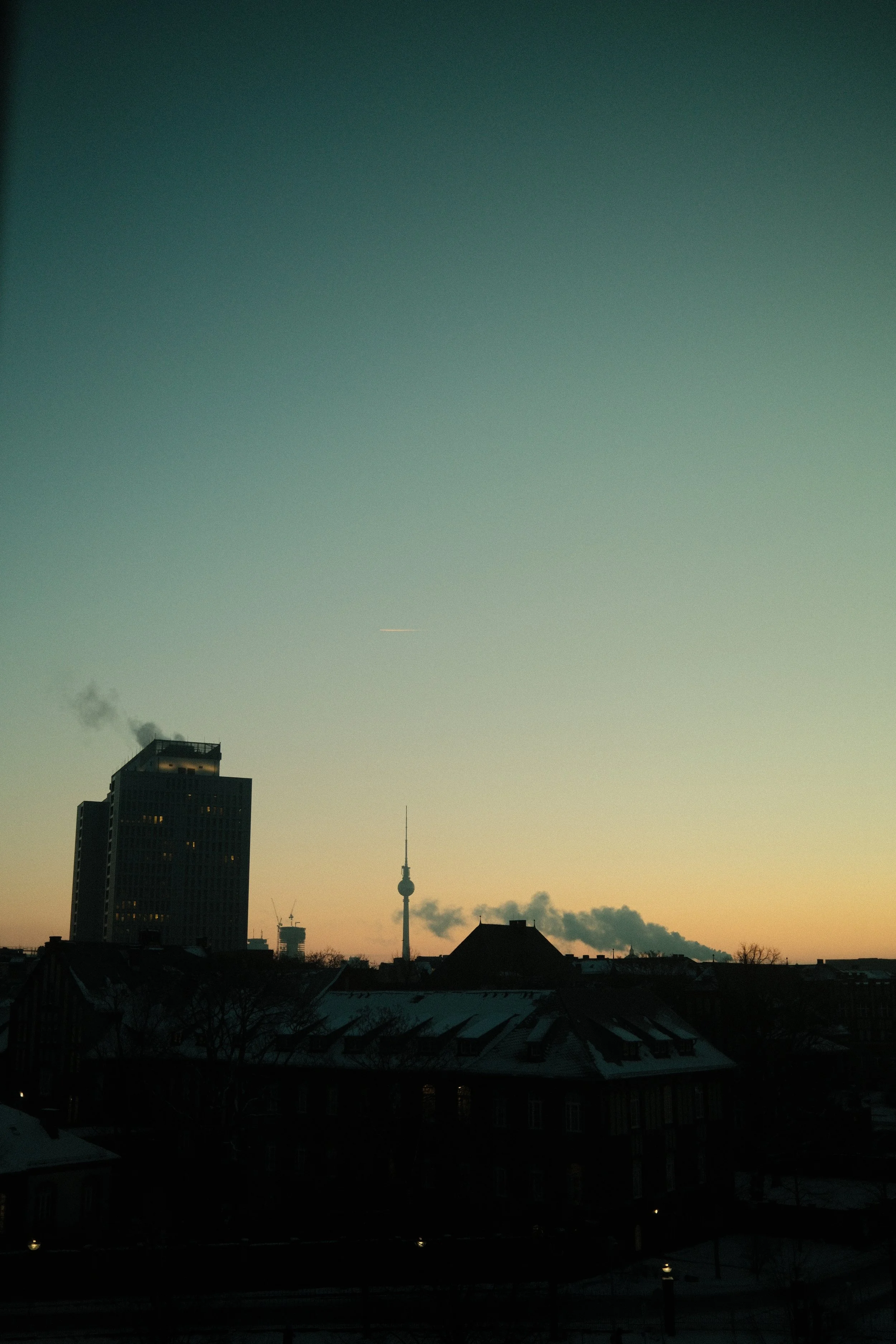 Stadtansicht bei Sonnenuntergang mit dunklen Gebäuden im Vordergrund und einem Fernsehturm in der Mitte im Hintergrund.