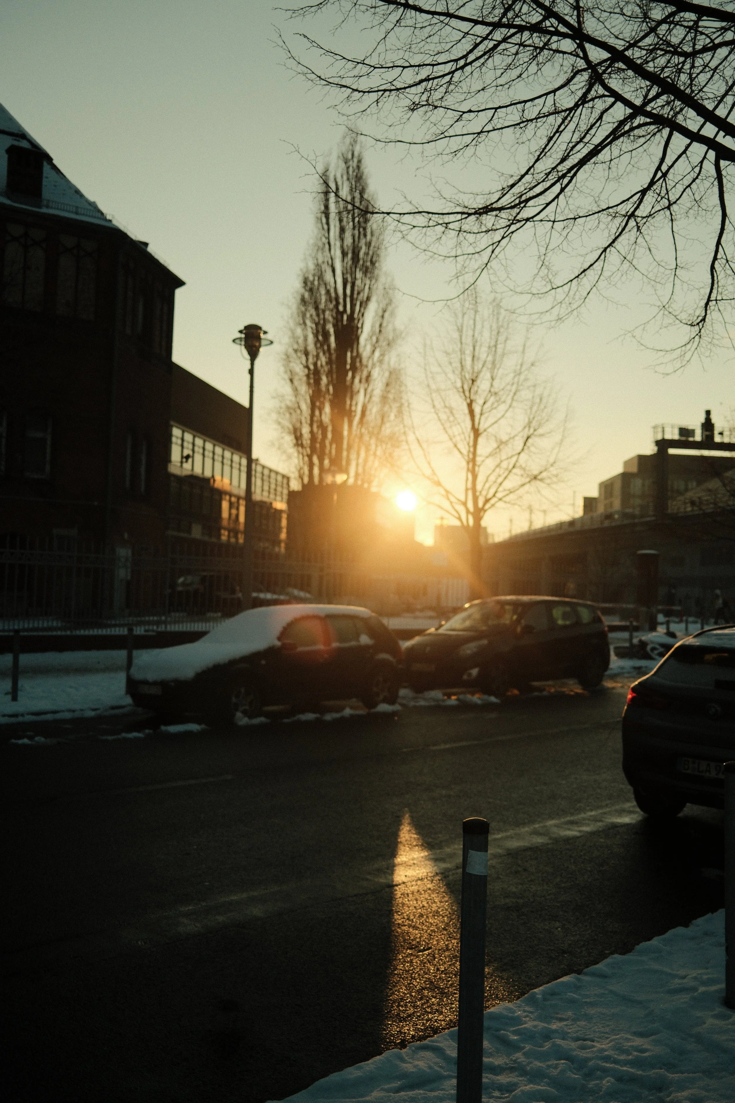 Die Sonne steht niedrig am Horizont, während Fahrzeuge an einem verschneiten Straßenrand parken. Es sind kahle Bäume und Gebäude im Hintergrund sichtbar, die in warmes Sonnenlicht getaucht sind.