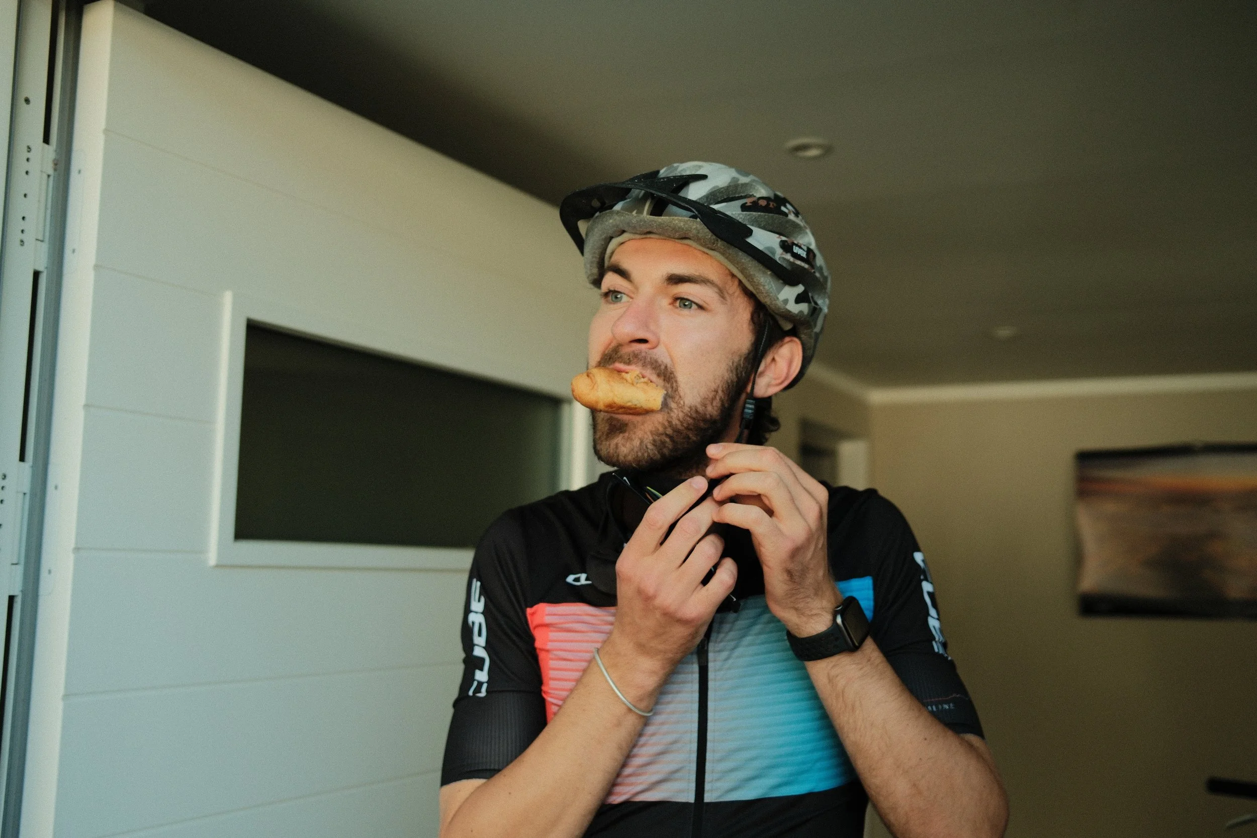 Ein Mann mit Fahrradhelm, der draußen ein Stück Gebäck im Mund hält und bei einer Radtour ist.